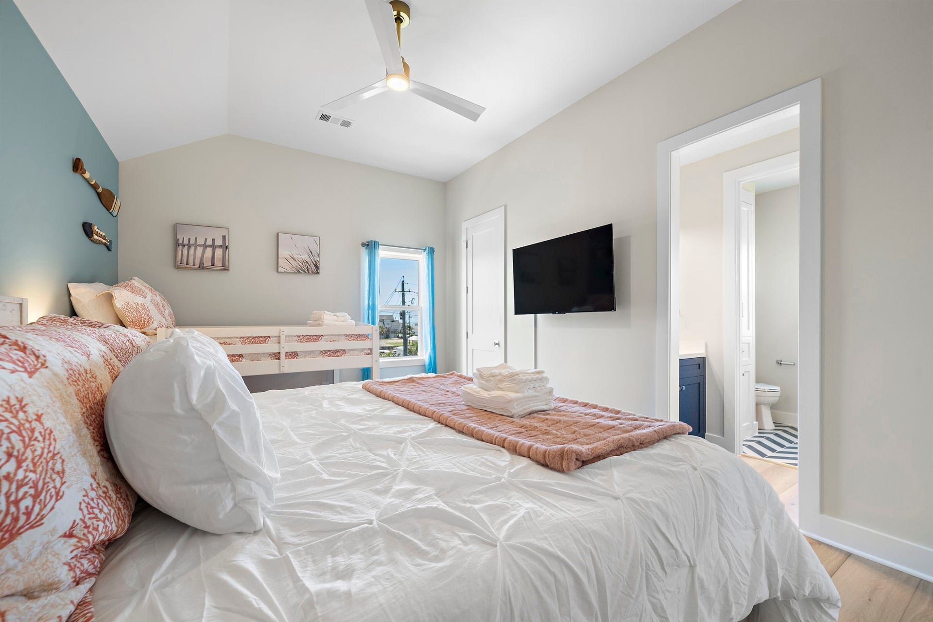 Bright and airy bedroom featuring a comfortable bed with coral-patterned pillows, a wall-mounted TV, and an en-suite bathroom. Perfect for relaxation and comfort.