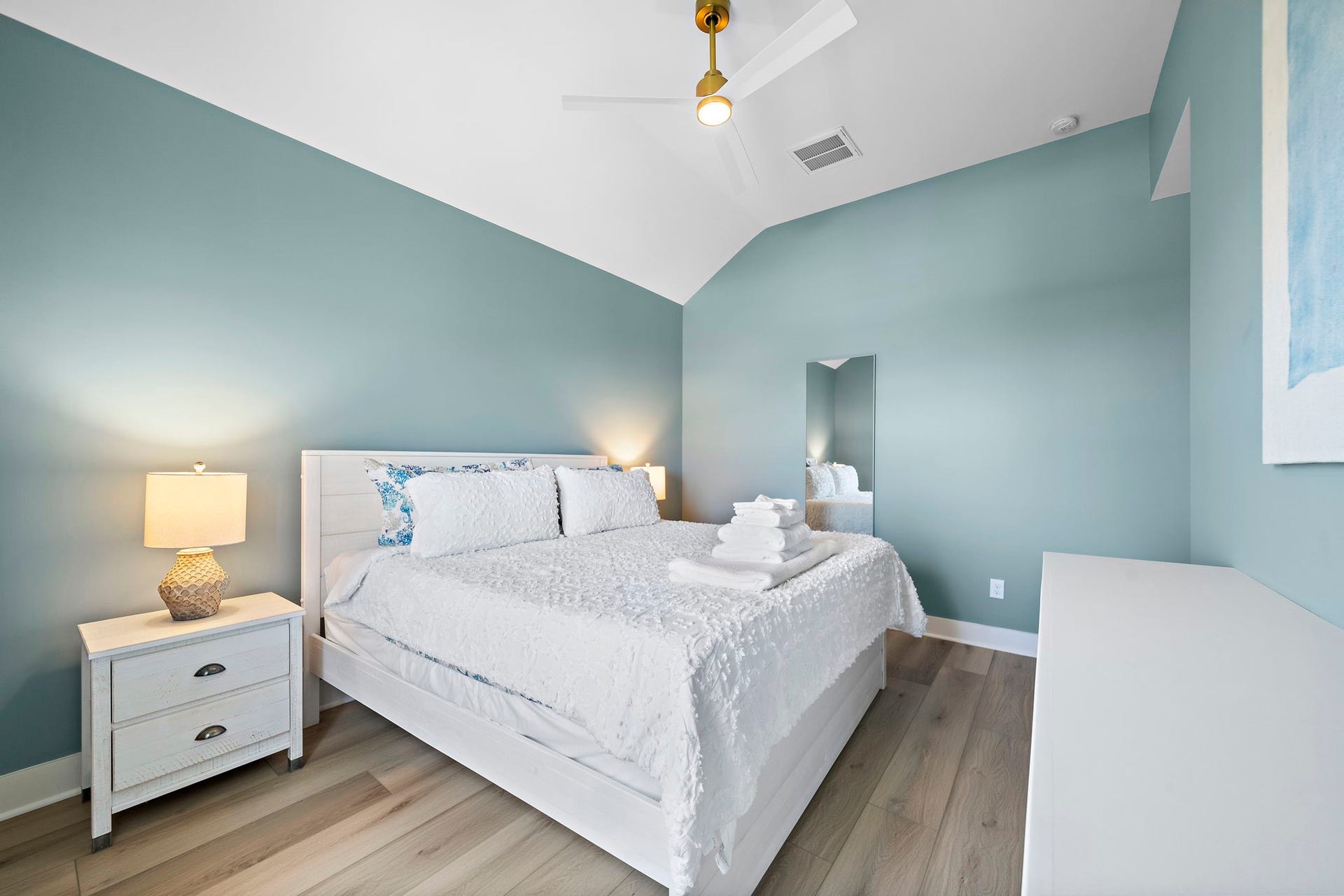 Serene bedroom with soothing blue walls, vaulted ceiling, and elegant wood flooring, featuring a cozy white bed and modern ceiling fan for ultimate comfort.
