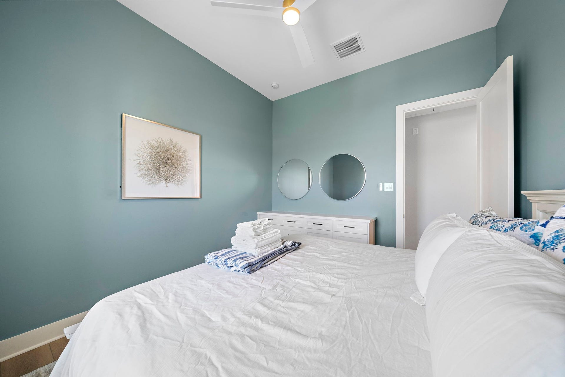 Serene bedroom with soothing blue walls, stylish artwork, and a cozy bed, complemented by a sleek dresser and circular mirrors.