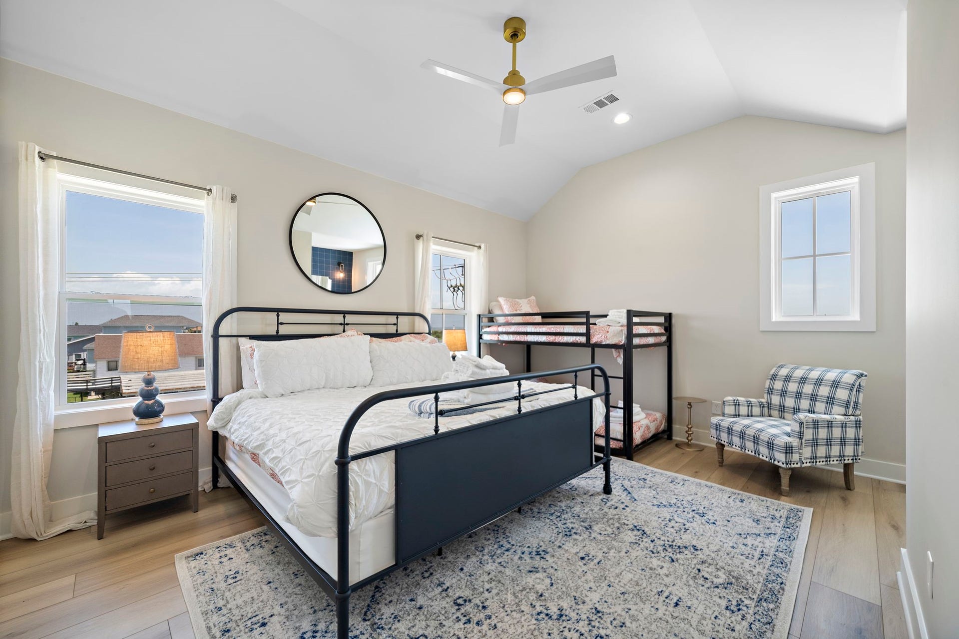 Bright and inviting bedroom featuring a stylish metal bed, cozy bunk beds, and a chic plaid armchair, all enhanced by natural light from large windows.