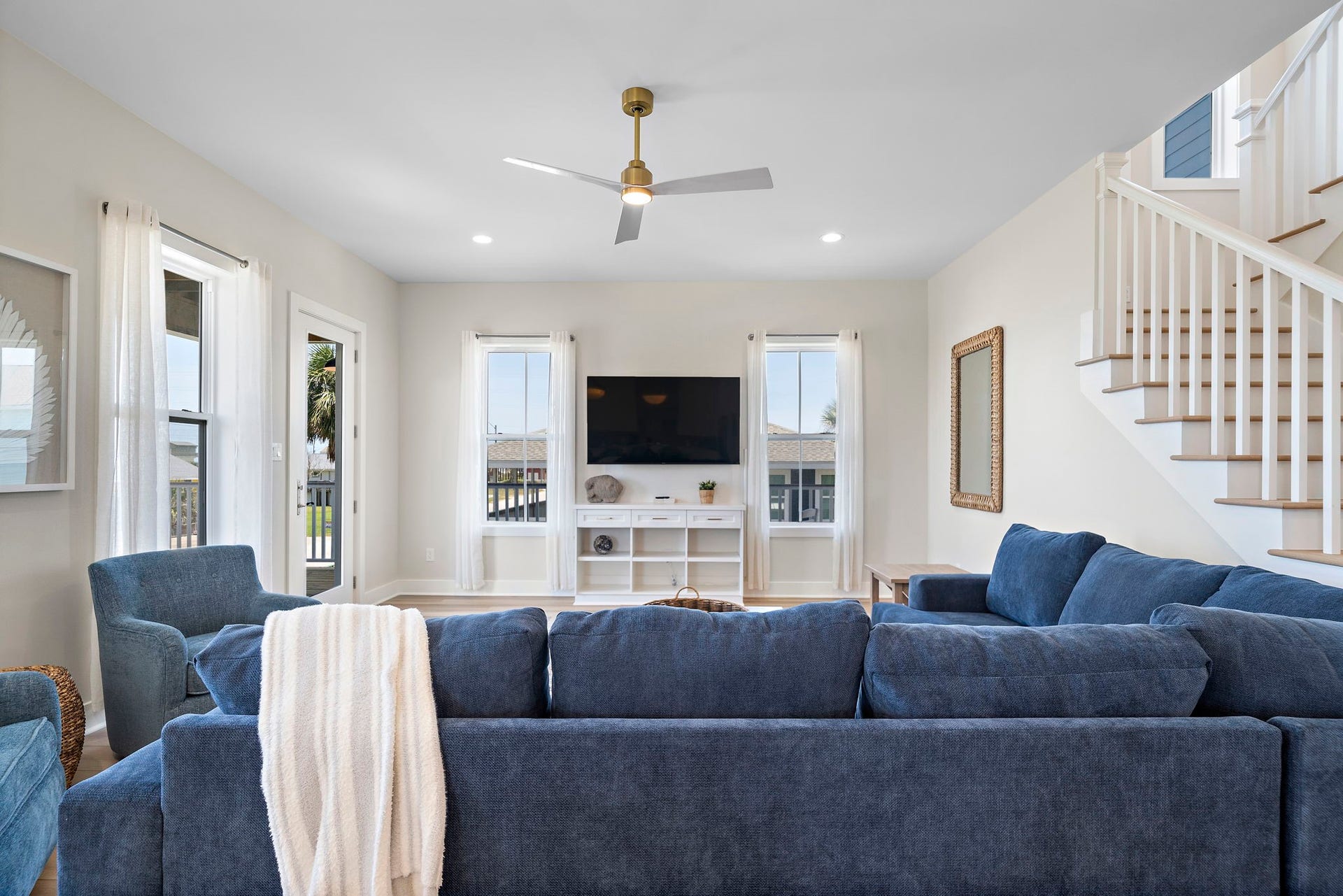 Spacious living room with abundant natural light, featuring a cozy blue sectional, ceiling fan, and modern entertainment center.