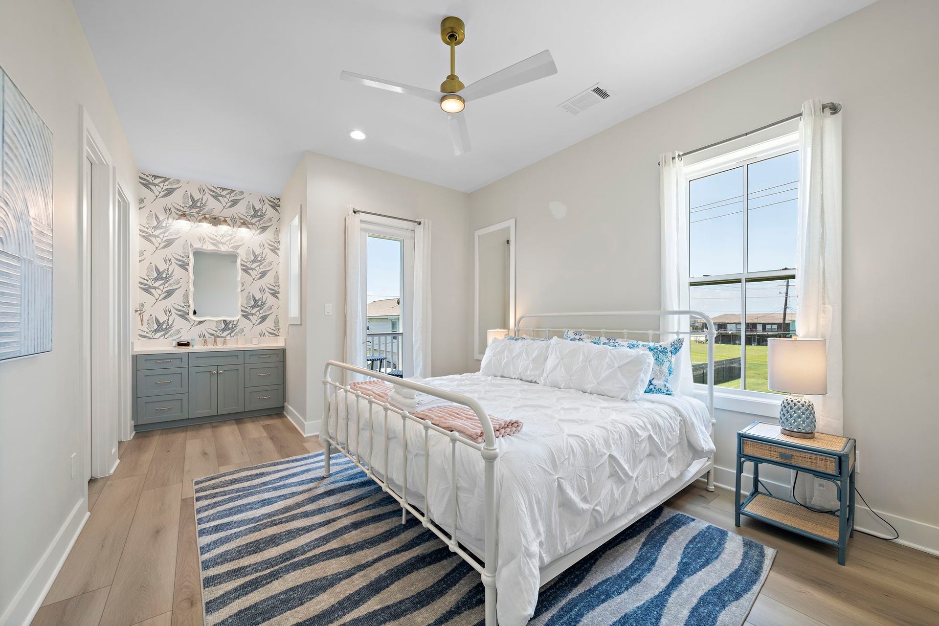 Bright and airy bedroom featuring a cozy bed, stylish ceiling fan, and en-suite bathroom with modern vanity and floral wallpaper. Large windows offer plenty of natural light.