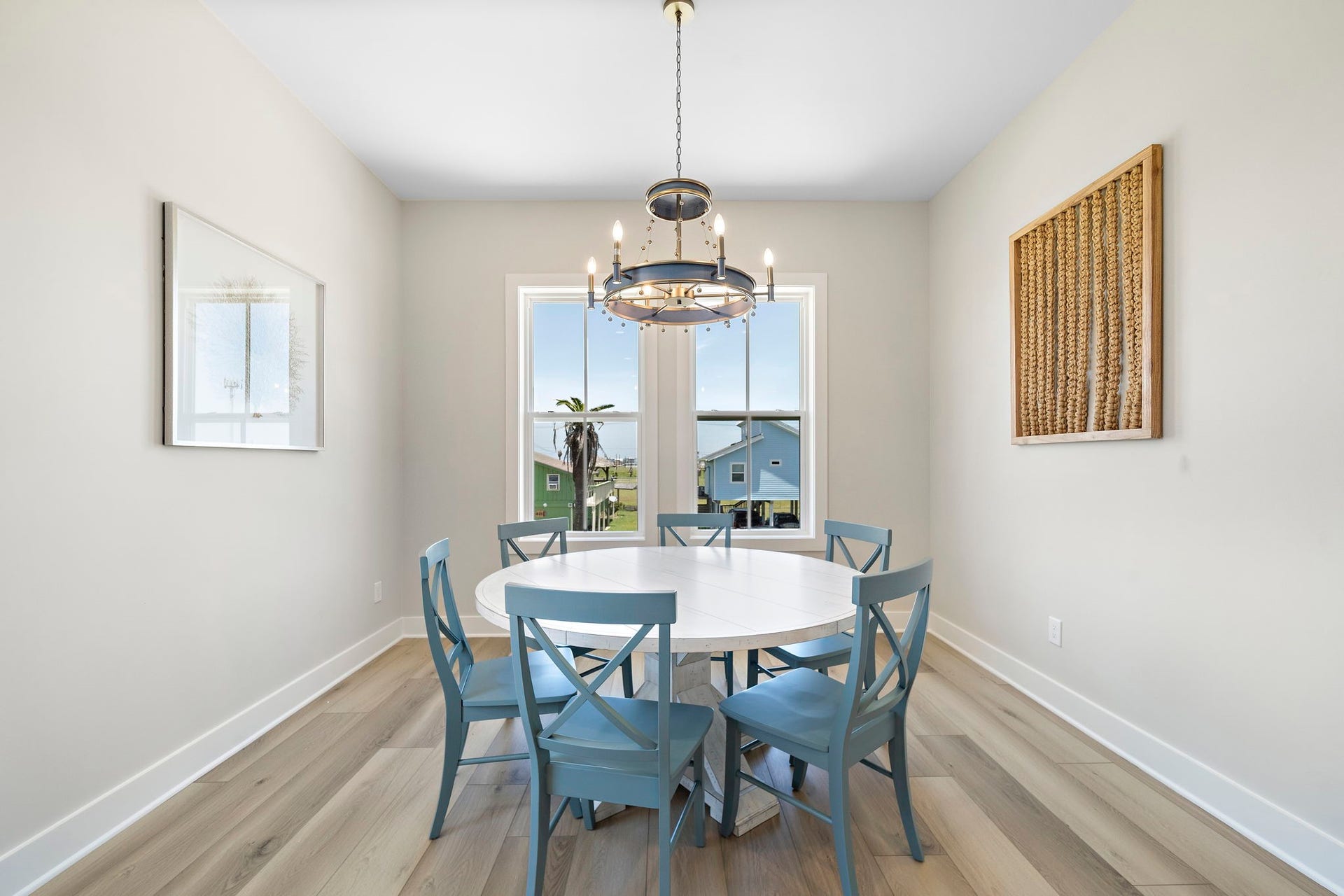 Bright dining area featuring a round table with blue chairs, complemented by a modern chandelier and large window offering scenic views.