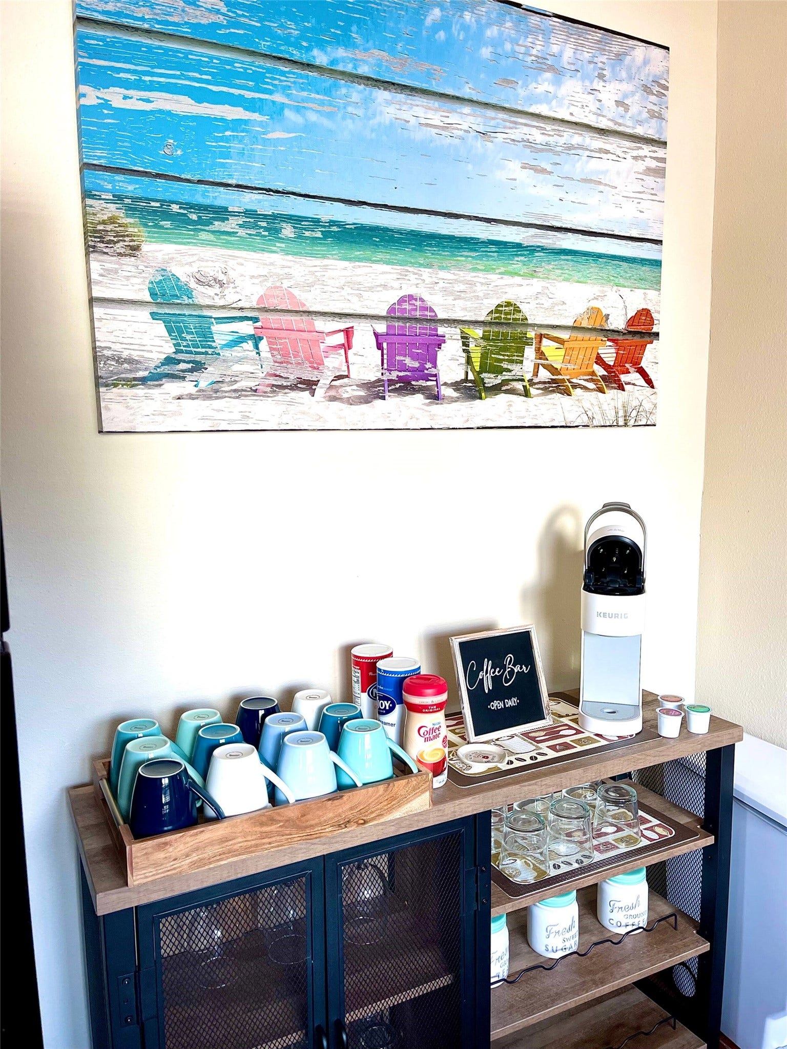 Charming coffee bar setup with colorful mugs, a variety of supplies, and a vibrant beach-themed artwork above, creating a cozy and inviting corner.