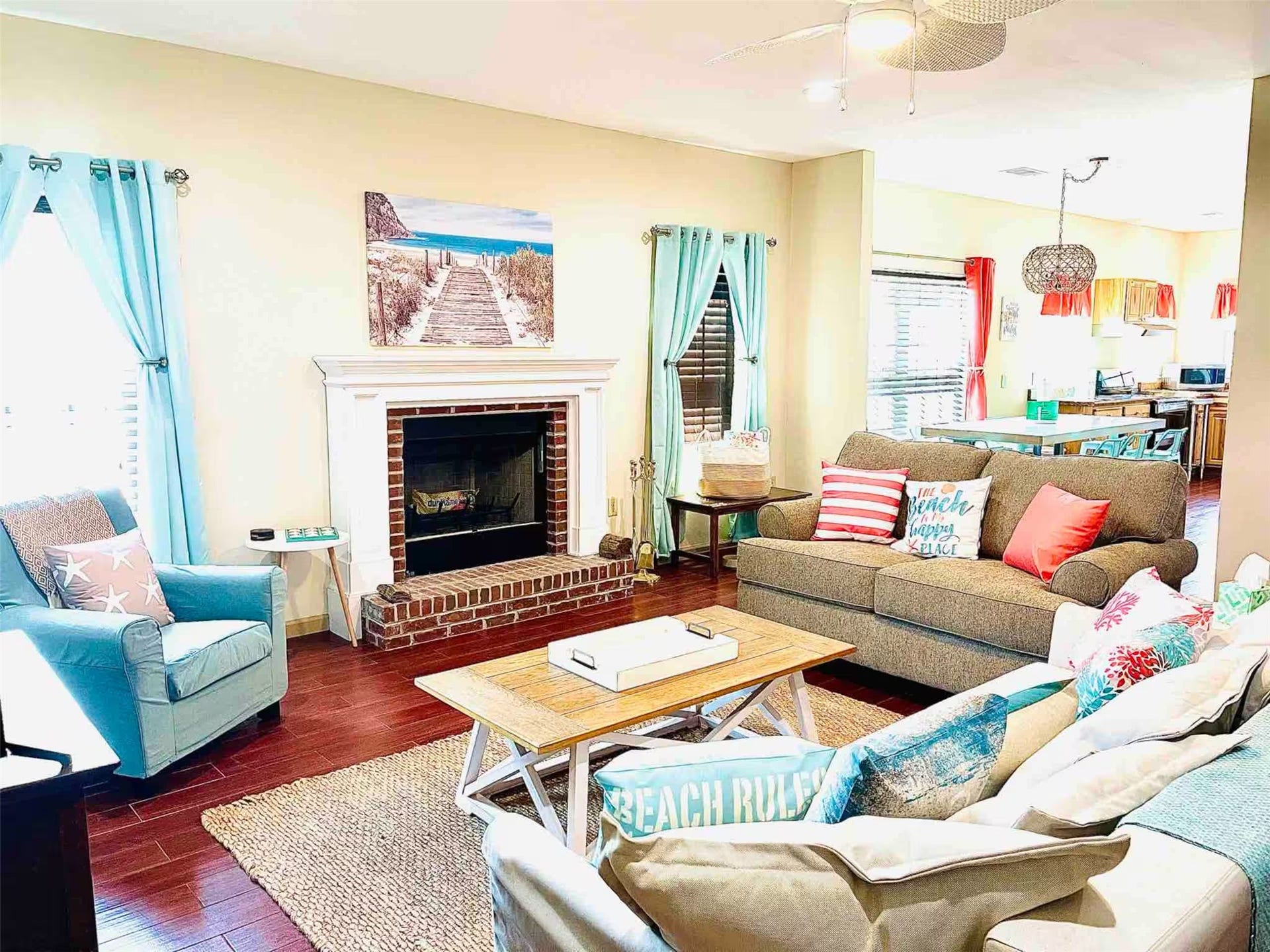 Charming living room with a cozy brick fireplace, bright turquoise curtains, and beach-themed decor, seamlessly flowing into the open kitchen area.