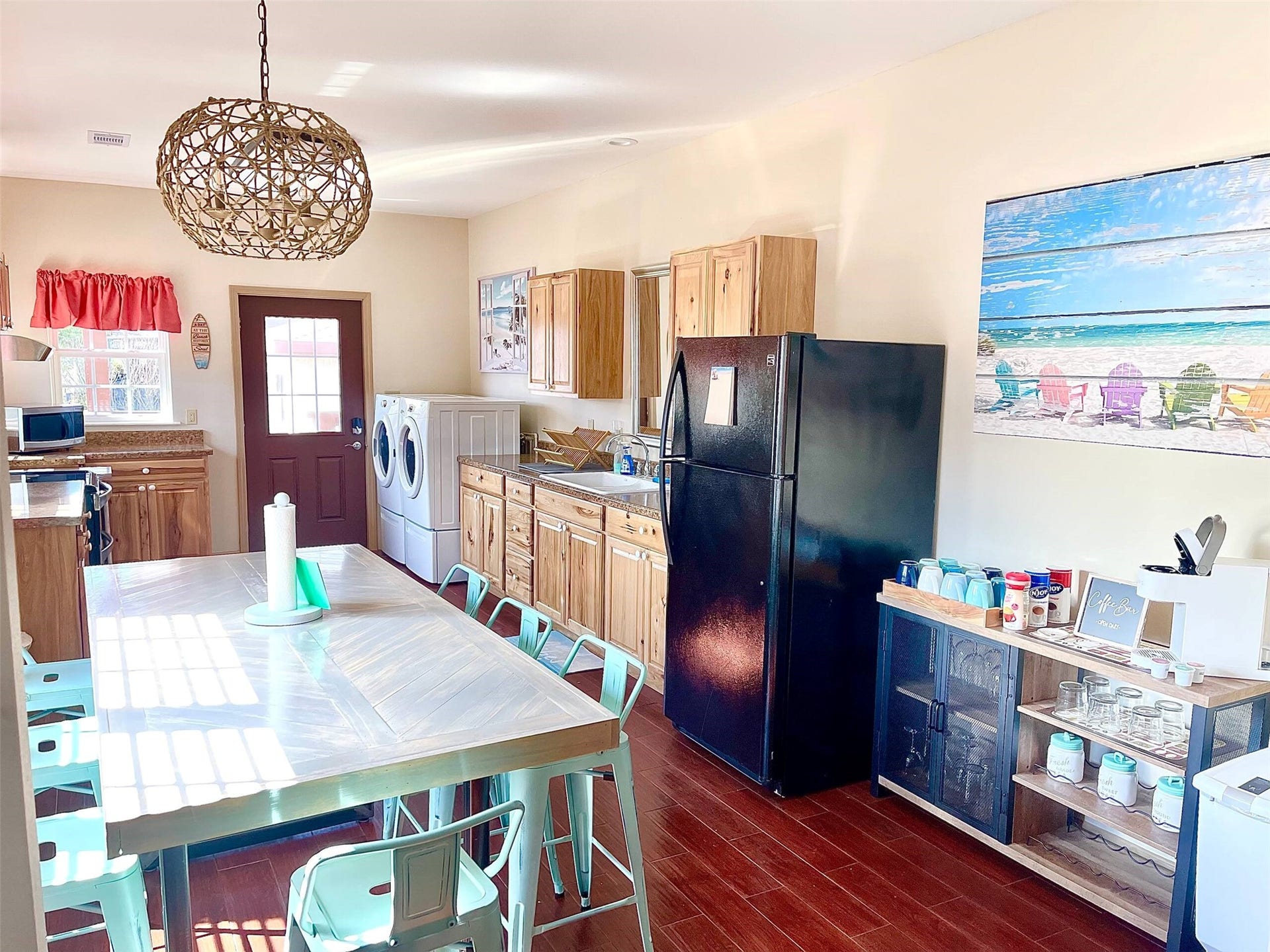 Charming kitchen with rustic wooden cabinetry, modern appliances, and a cozy dining area featuring teal chairs. Bright and inviting, it also includes laundry facilities and vibrant beach-themed decor.