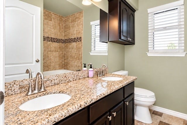 928 Oleander Street, Lake Jackson, TX 77566 Elegant bathroom featuring a dual-sink granite countertop, rich dark cabinetry, and a tiled shower area with a decorative accent strip.