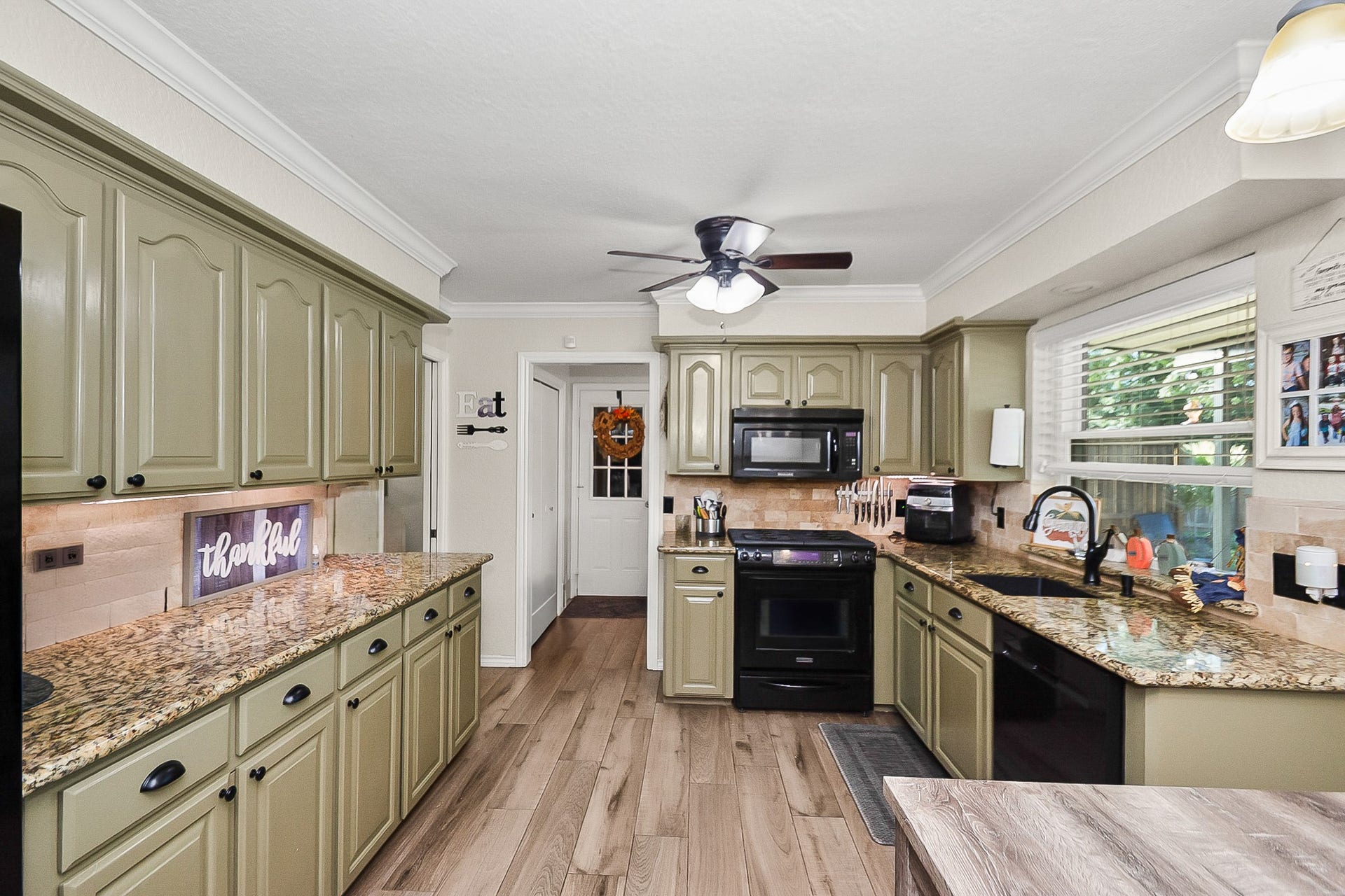 928 Oleander Street, Lake Jackson, TX 77566 Charming kitchen with olive green cabinetry, sleek granite countertops, and modern black appliances. Natural light streams through the large window, highlighting the spacious layout and warm wood look flooring.
