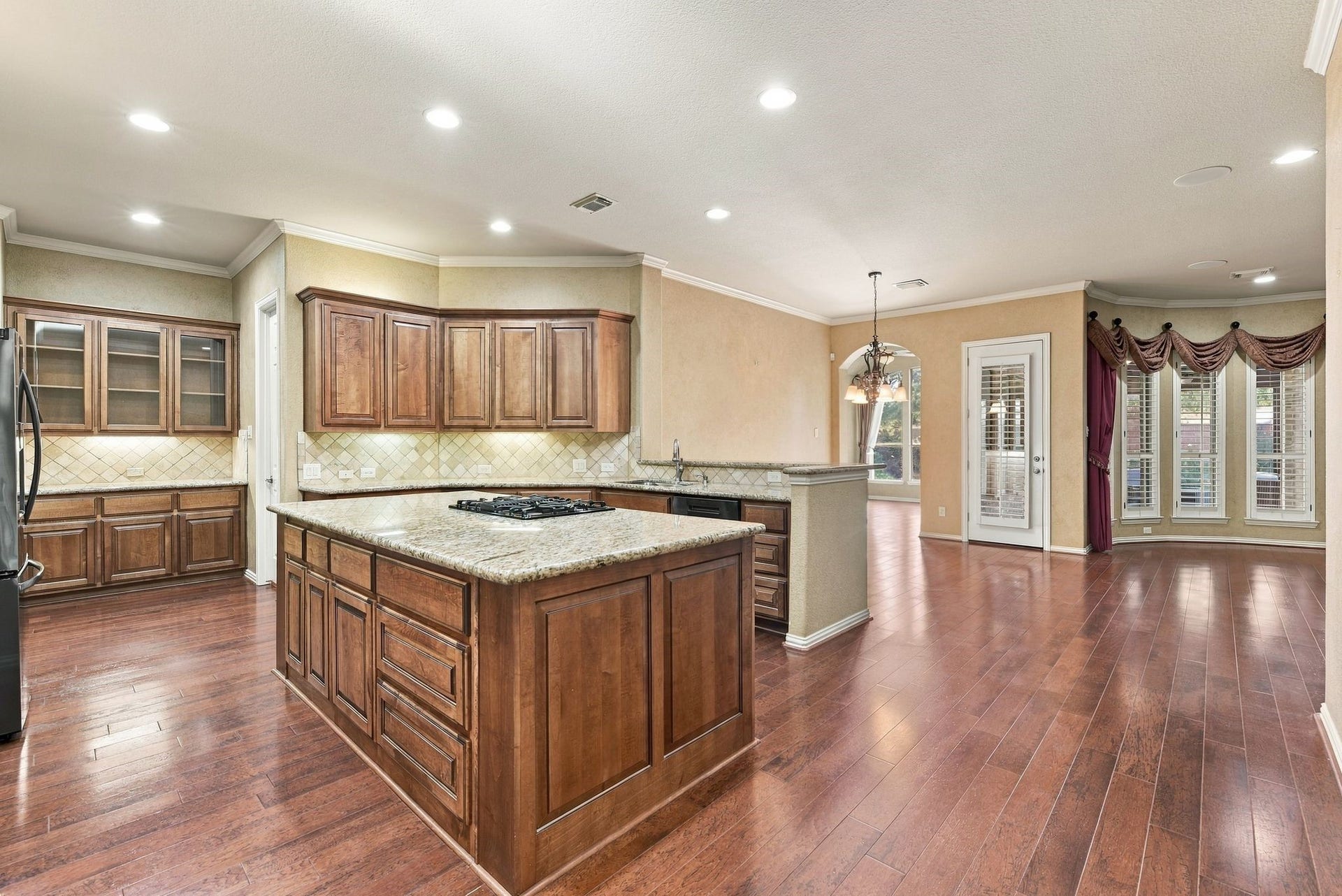 Spacious kitchen with rich wooden cabinetry, granite countertops, and a central island with a gas cooktop. The open layout flows into the breakfast area.