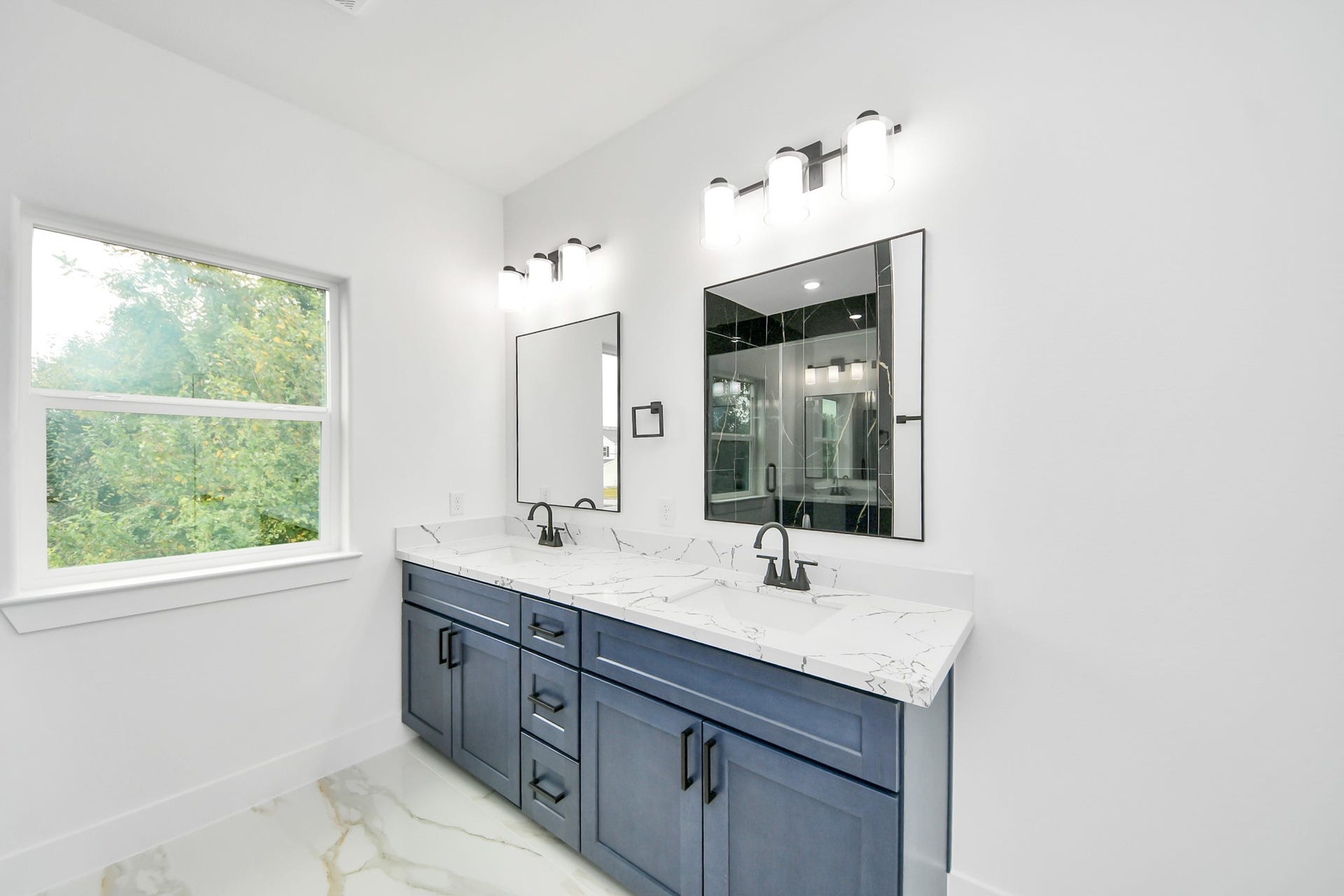 6707 Knox Street, Houston, TX 77091 Elegant bathroom featuring a double vanity with sleek blue cabinetry, polished marble countertops, and modern fixtures, complemented by ample natural light from the large window.