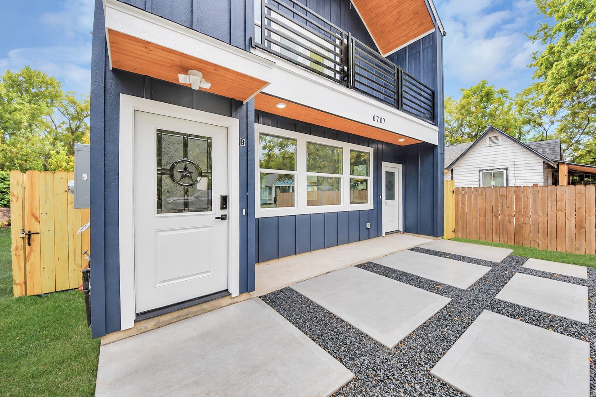 6707 Knox Street, Houston, TX 77091 Stylish modern home with a striking navy exterior, accented by white trim and wood paneling. Features include a spacious porch, large windows, and a sleek balcony. The landscaped yard and unique concrete and stone pathway enhance the inviting entrance.