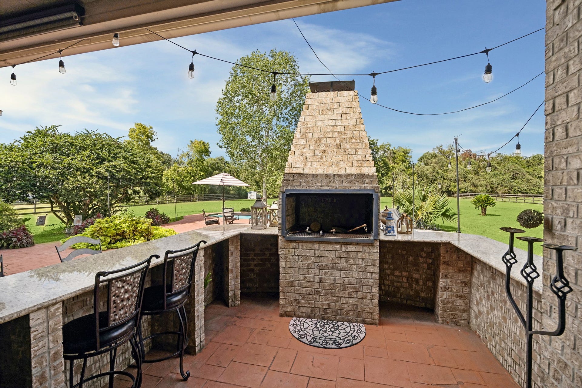 1319 Brazoswood Place, Richmond, TX 77406 Enjoy outdoor entertaining on the covered patio, boasts a brick fireplace, wraparound stone countertops and bar seating. String lights add ambiance, while views of the pool, expansive lawn and mature trees create a picturesque setting.