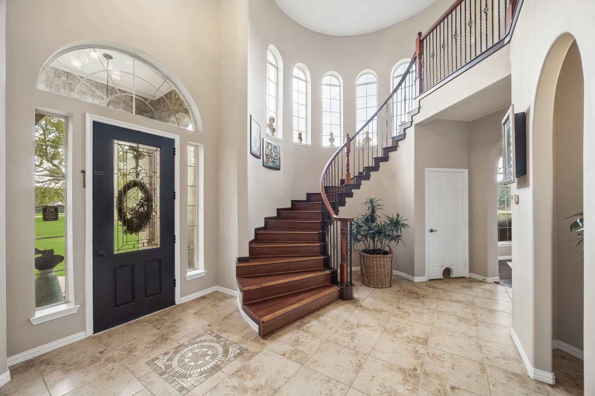 1319 Brazoswood Place, Richmond, TX 77406 A grand, light-filled foyer showcases a dramatic curved hardwood staircase with wrought-iron railings, and soaring windows. A decorative glass-paneled entry door and detailed tile inlay add warmth and character to the impressive two-story entrance.
