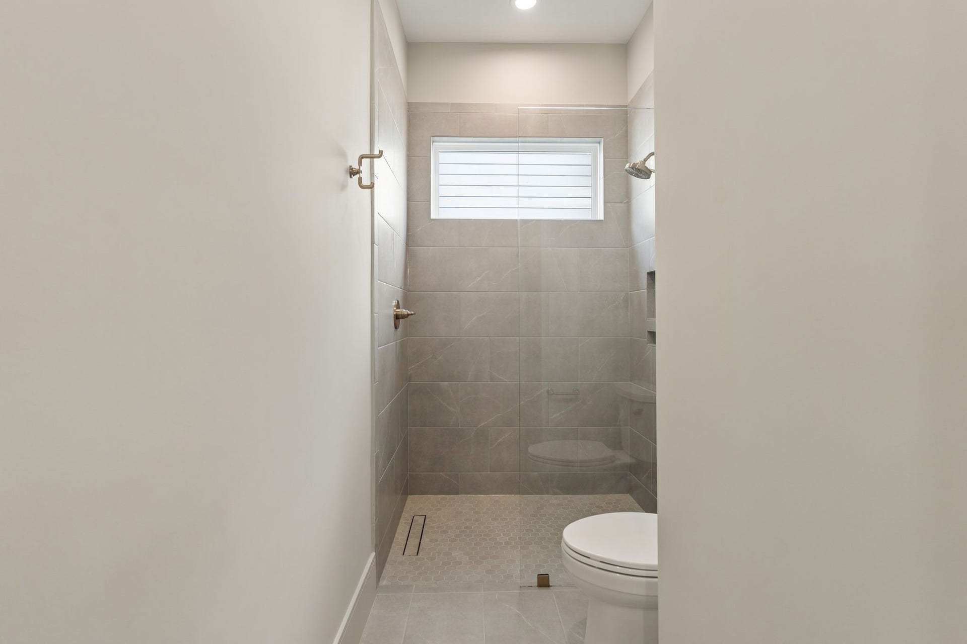 711 E 12th Street, Houston, TX 77008 Soft gray tile, warm brass fixtures, and a frameless glass shower give this bath a refined, understated feel. A high window brings in natural light, while hexagonal shower tile and a recessed niche add thoughtful detail.