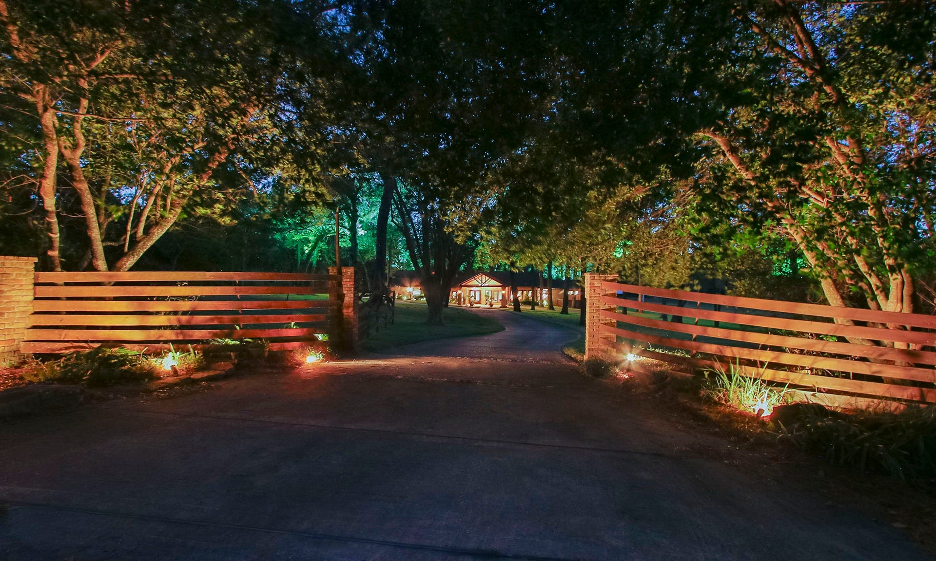23107 Kobs Road, Tomball, TX 77377 This photo showcases a welcoming driveway with a wooden gate, lined with trees and soft landscape lighting leading to a cozy, well-lit home nestled in a serene, wooded setting.
