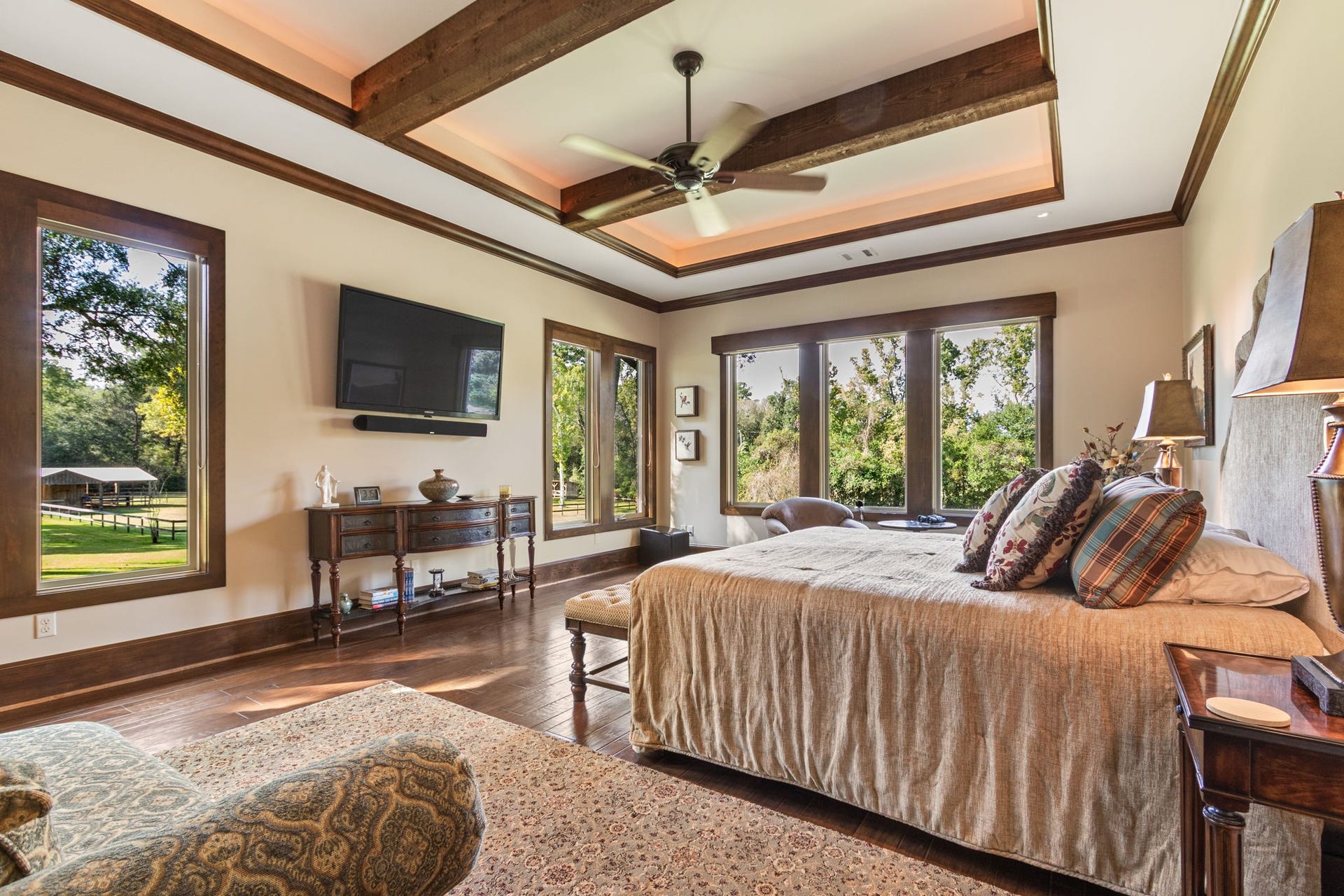 23107 Kobs Road, Tomball, TX 77377 This spacious primary suite features large windows with scenic views, wood-beamed ceilings, and elegant hardwood floors. A ceiling fan provides comfort, while creating a perfect blend of luxury and relaxation.