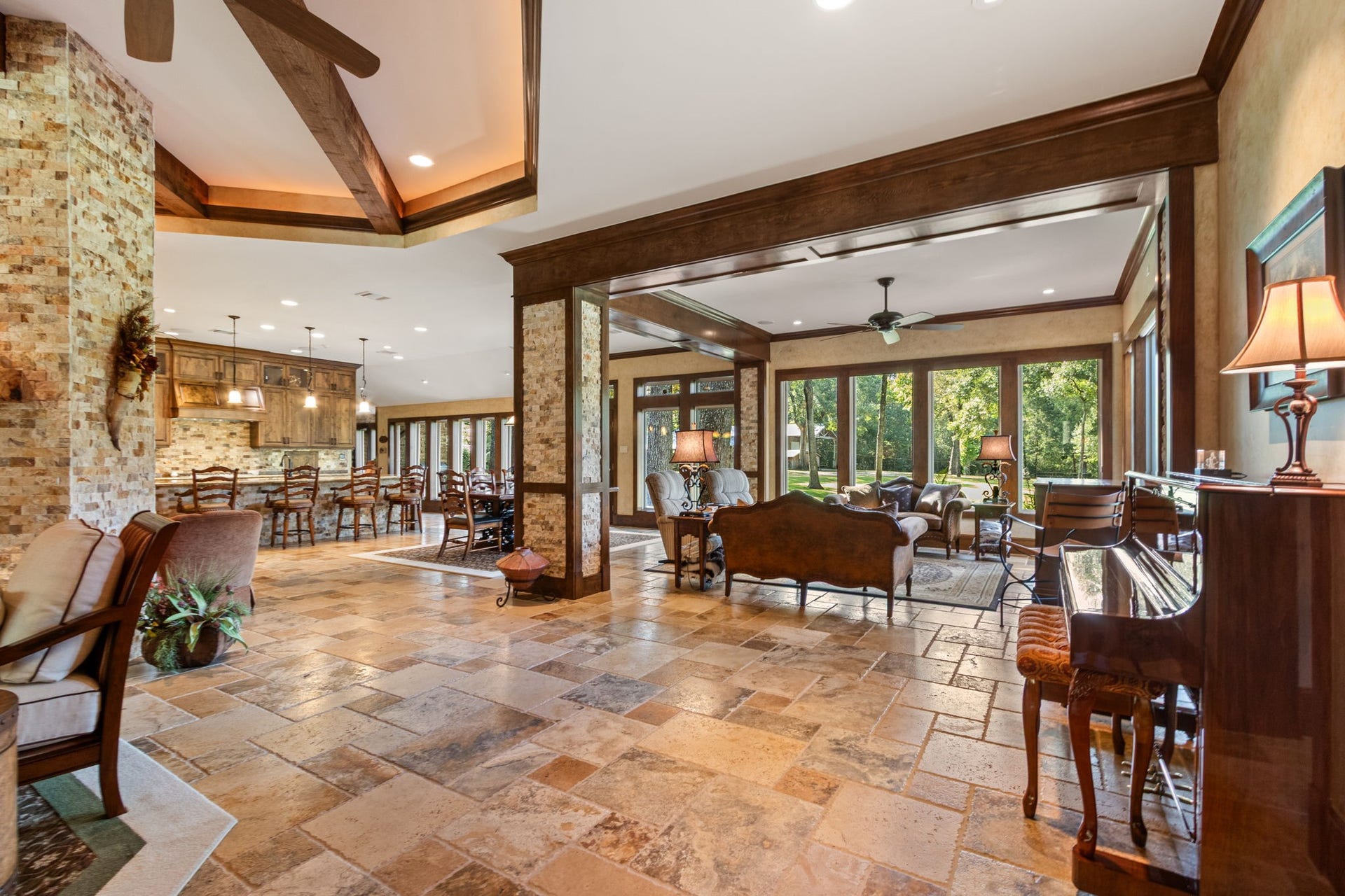 23107 Kobs Road, Tomball, TX 77377 This spacious, elegant home features an open floor plan with a warm, earthy color palette. It includes a cozy seating area, large dining space, and ample natural light from expansive windows overlooking lush greenery. The rich wood accents and travertine flooring add to its inviting charm.