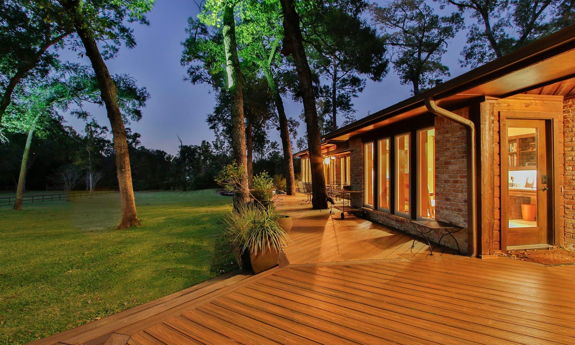 23107 Kobs Road, Tomball, TX 77377 This photo showcases a charming home with a warm, wooden deck and large windows, set against a serene, wooded backdrop. A spacious deck wraps around the house, perfect for relaxing or entertaining, offering views of the expansive lawn and tall trees.