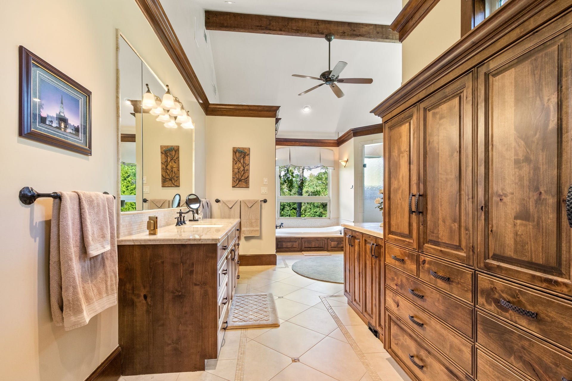 23107 Kobs Road, Tomball, TX 77377 This spacious primary bathroom features elegant wood cabinetry and a double vanity. The room is well-lit with modern fixtures and showcases a warm, inviting design with tile flooring and a ceiling fan. Perfect for relaxation and functionality.