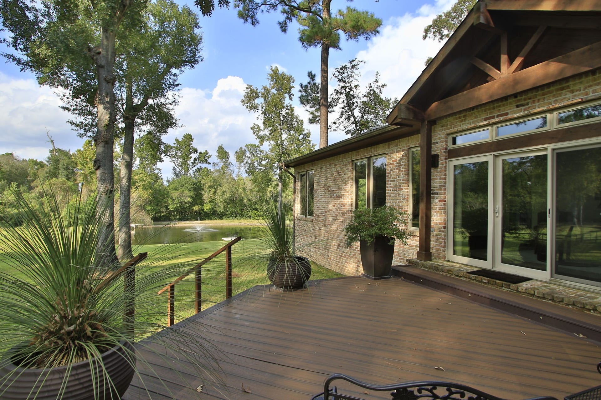 23107 Kobs Road, Tomball, TX 77377 This photo shows a serene backyard view from a wooden deck, featuring a brick house with large windows and sliding glass doors. The deck overlooks a lush lawn and a small pond with a fountain, surrounded by trees, creating a peaceful and private outdoor space.