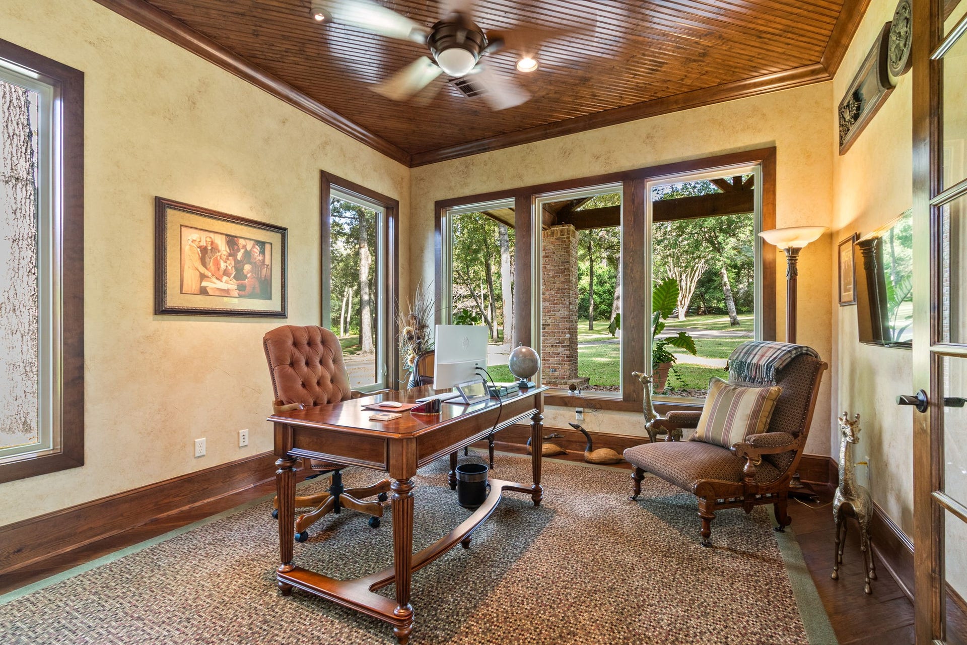 23107 Kobs Road, Tomball, TX 77377 This home office features large windows with a view of lush greenery, providing ample natural light. It includes a beaded board ceiling, wood floors, ceiling fan and warm lighting.