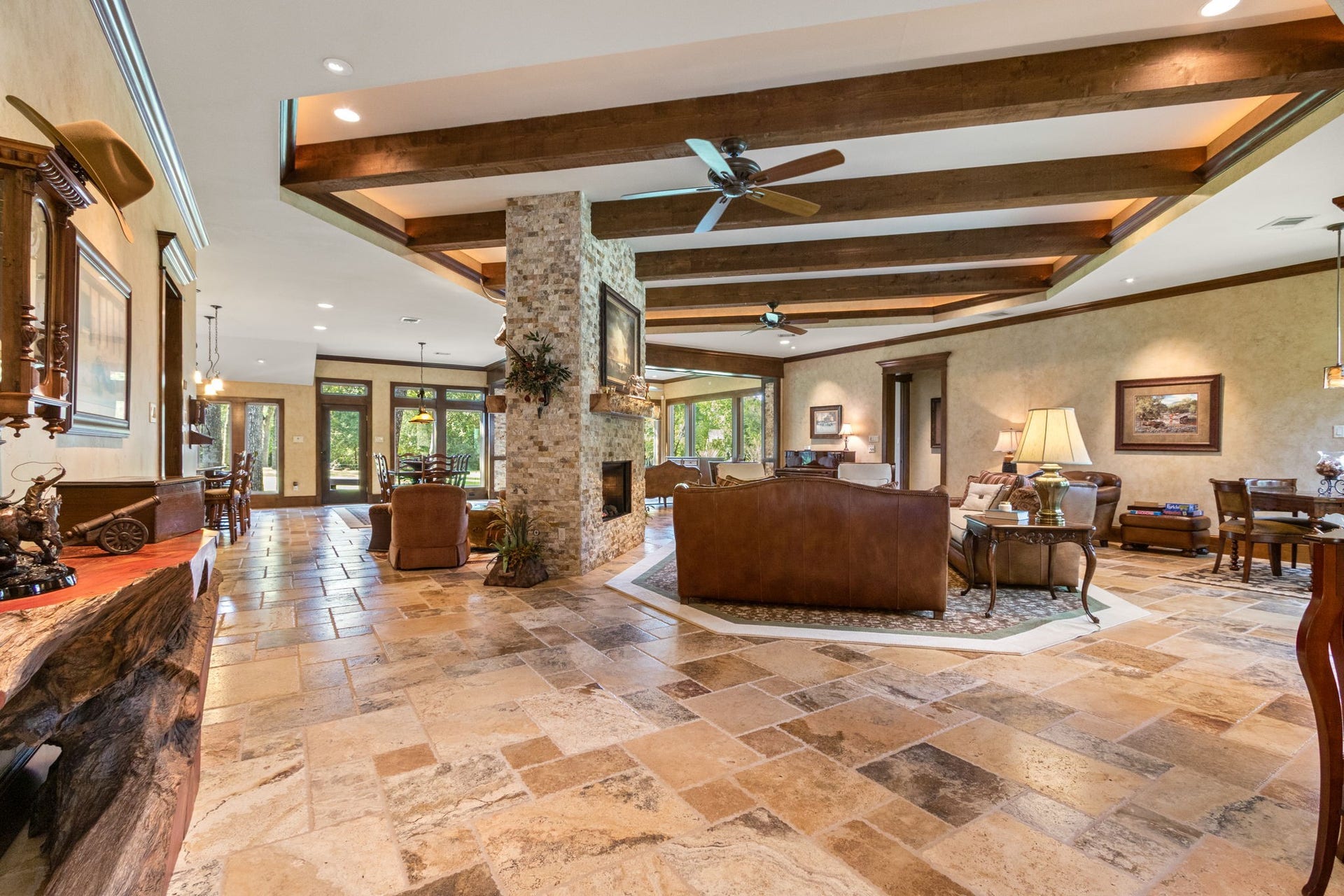 23107 Kobs Road, Tomball, TX 77377 This spacious, open-concept living area features a rustic stone fireplace, exposed wooden beams, and large windows that flood the space with natural light. The elegant travertine tile flooring complements the cozy family spaces, while the adjoining dining space offers a seamless flow for entertaining.