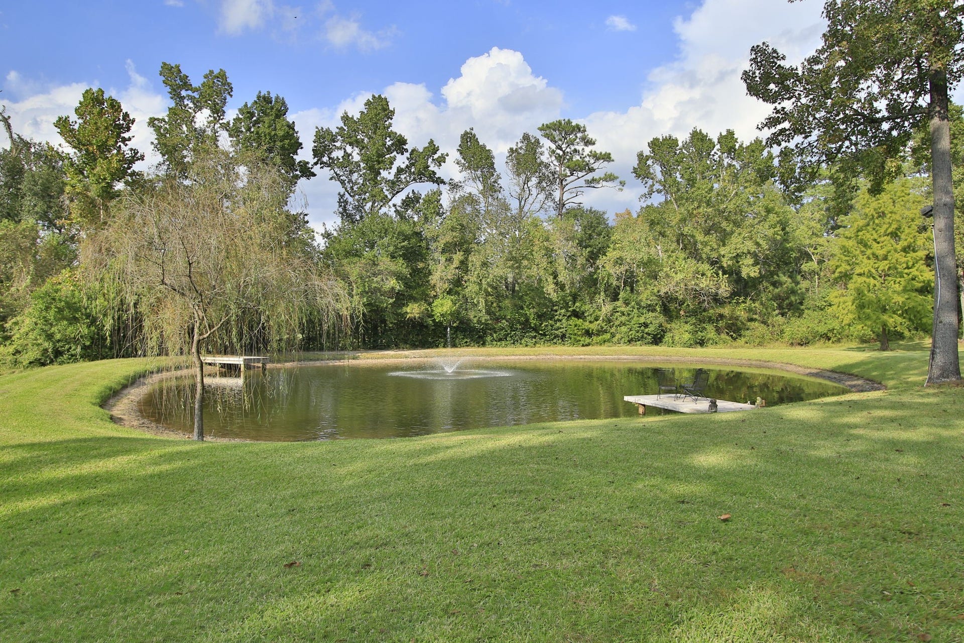 23107 Kobs Road, Tomball, TX 77377 This photo showcases a serene backyard with a pond featuring a central fountain. The area is surrounded by lush trees and well-maintained grassy landscaping, offering a peaceful and private outdoor space.