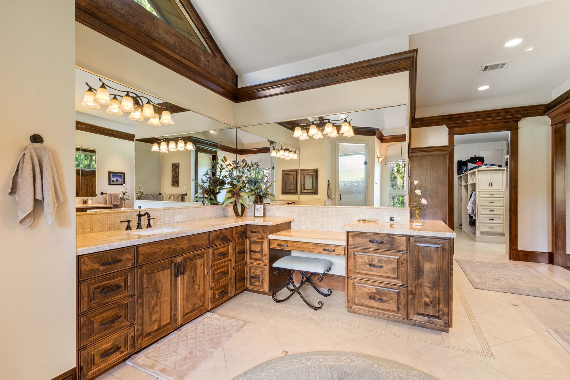 23107 Kobs Road, Tomball, TX 77377 This photo showcases a spacious and elegant primary bathroom with rich wood cabinetry, a large vanity area, and ample lighting. The room features a wide mirror, a comfortable seating area for your vanity bench, and an adjacent walk-in closet, providing both functionality and luxury.