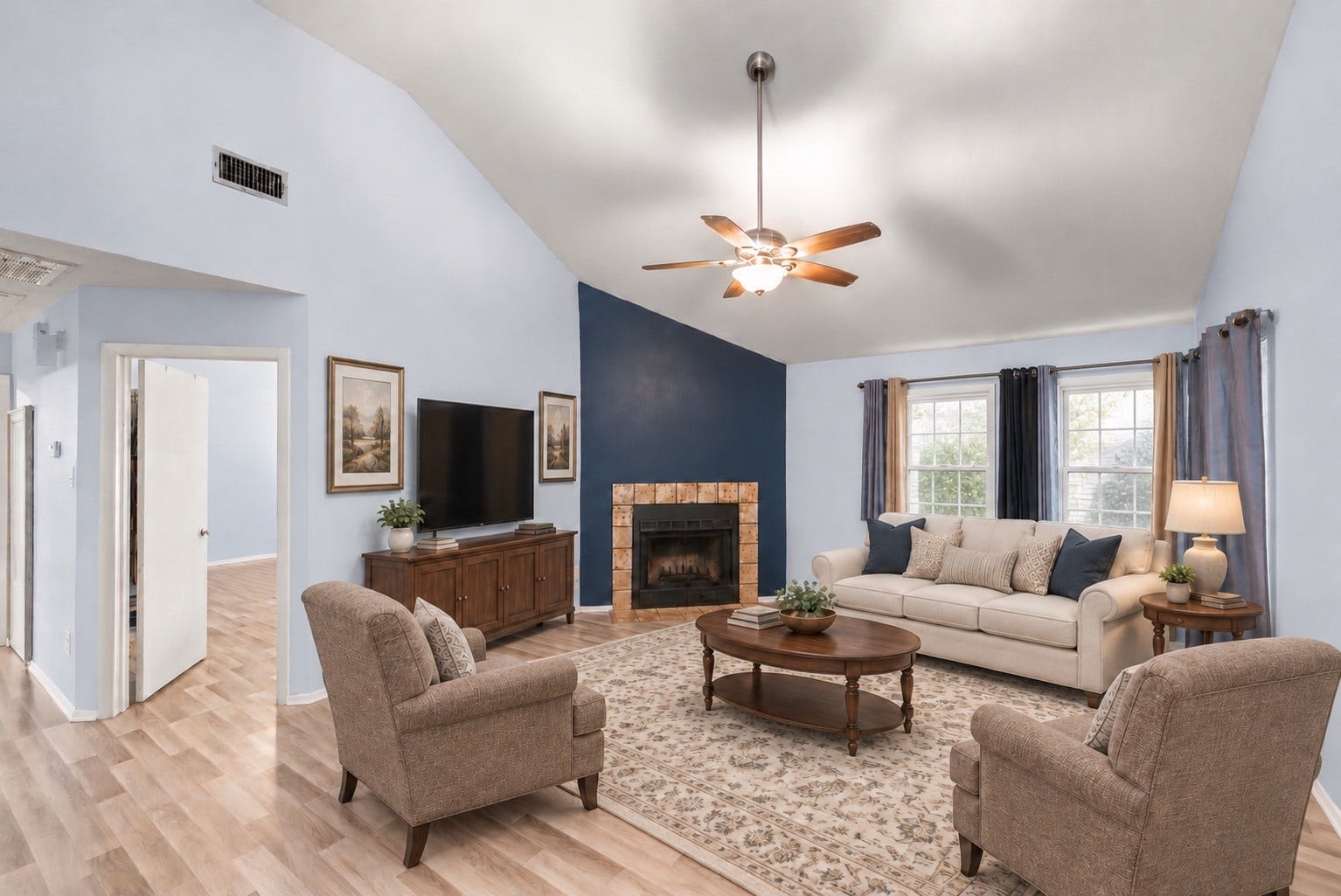 A full sofa, two armchairs and a media console fit easily in the 17'9" x 14'8" Living Room. The corner fireplace with its tile surround sits against a navy accent wall, and a ceiling fan with light hangs from the vaulted ceiling. Two curtained windows bring in daylight. The doorway on the left opens to the bedroom hall, and the Primary Bedroom door sits just off the Living Room. The fireplace stays in view from the Dining Area as well. Virtually staged.