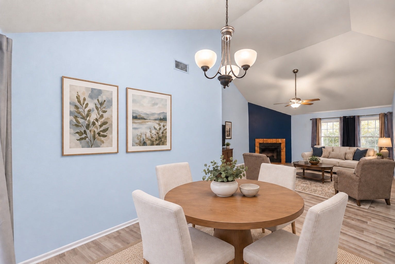 From the Dining Area, the view runs straight across the Living Room to the fireplace and windows. A round table with four chairs fits under the hanging light fixture with room to walk around it. The long blank wall has space for artwork or a buffet. Wood-look floors connect the two rooms and keep the table part of the main living space. The Kitchen is just around the corner for serving, and the sliding glass door is behind the table. Virtually staged.
