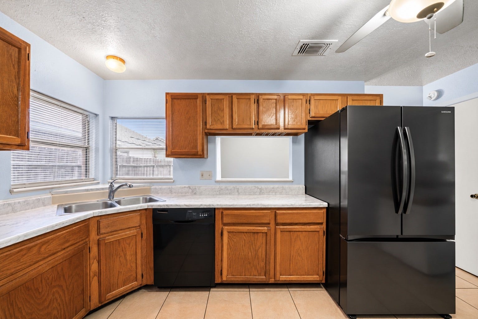 Two windows sit above the double sink in the corner, with counters running along both walls for meal prep. The included Samsung French-door refrigerator stands at the end of the counter run, and the black dishwasher is built in next to the sink. Upper and lower wood cabinets give plenty of storage, and the light marble-look counters are easy to wipe down. A wall opening above the counter looks into the Dining Area, keeping the cook connected to the rest of the main living space.