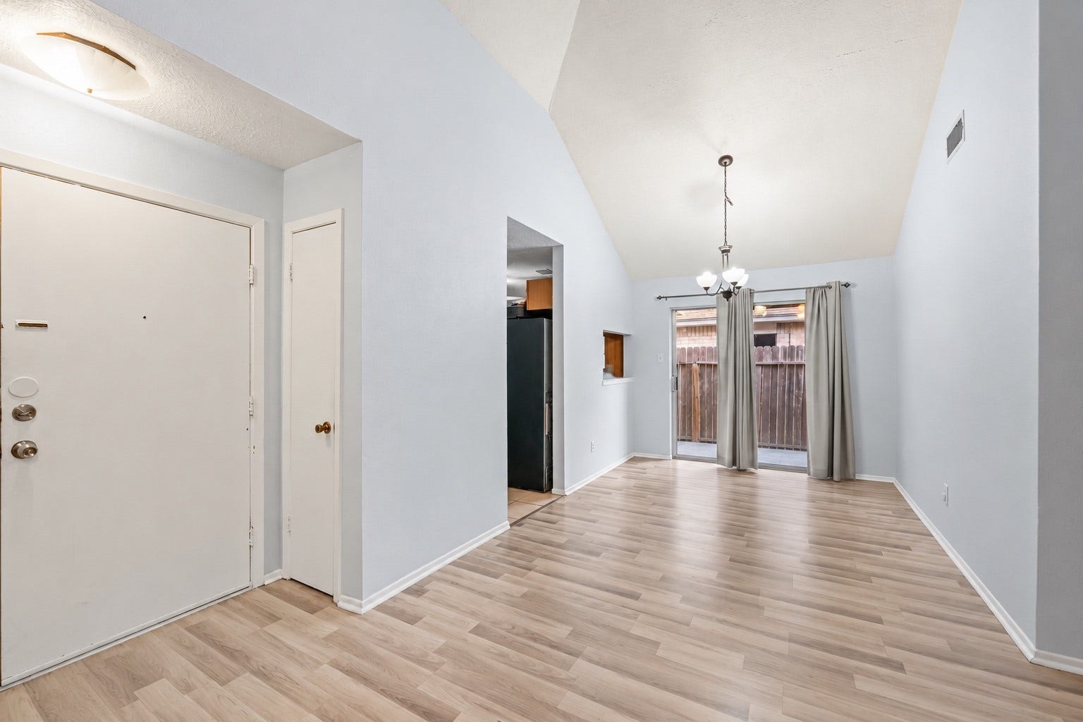 Standing at the front door with the room empty, the layout is easy to read: Dining Area straight ahead, Kitchen through the doorway on the left and the Living Room to the right. The vaulted ceiling and light walls make the main living space feel larger than its footprint. Daylight comes in through the sliding glass door from the backyard. A ceiling light hangs over the Foyer, and the coat closet keeps jackets out of the way when friends come over.