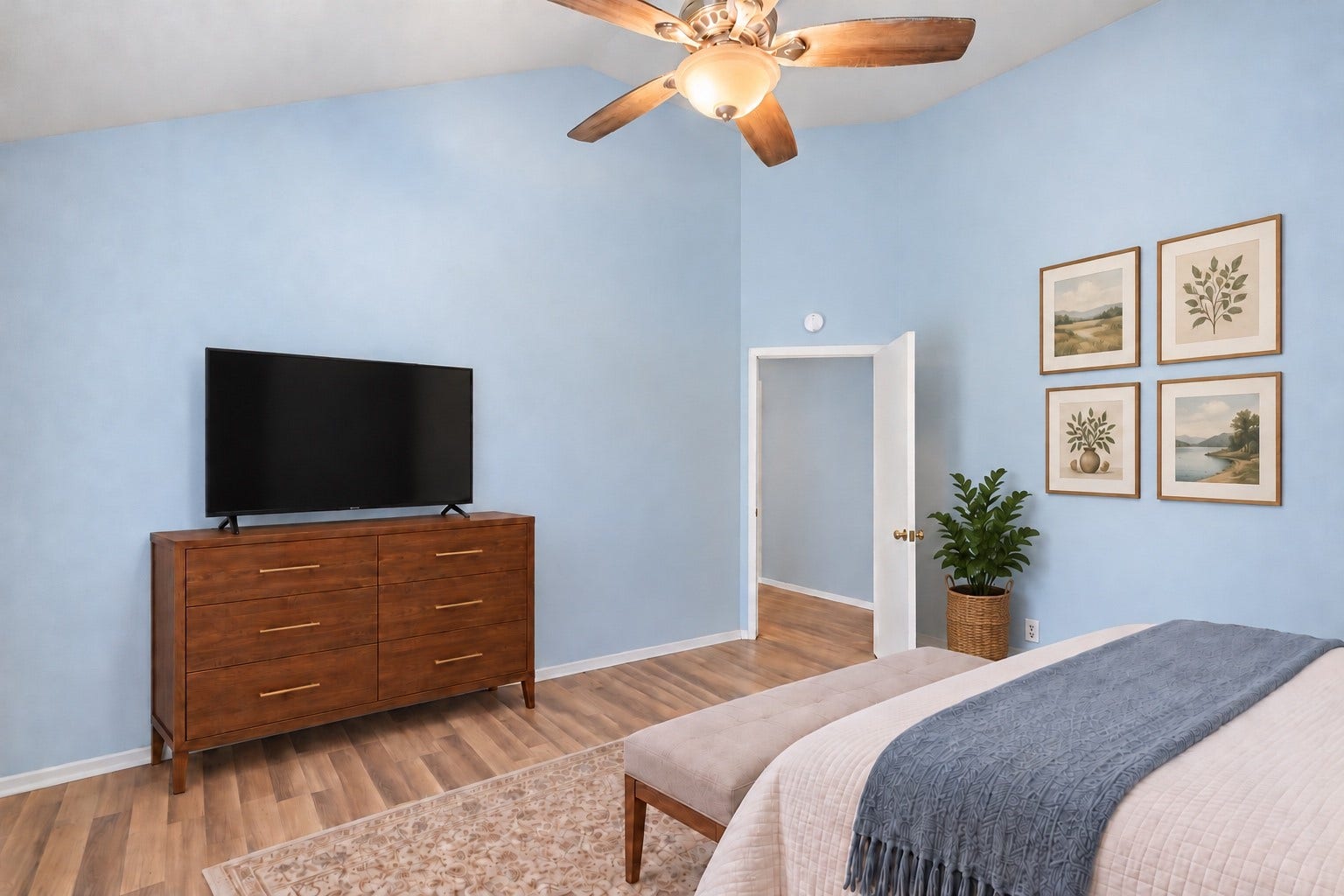 Turned toward the door, the Primary Bedroom has a full wall for a long dresser across from the bed. The angled entry door opens from the Living Room, keeping the room close to the main living space but apart from the other bedrooms down the hall. The ceiling fan with light hangs from the vaulted ceiling, and wood-look flooring runs to the doorway. Framed art fills the wall beside the doorway. A bench fits at the foot of the bed, with walkway room on both sides. Virtually staged.