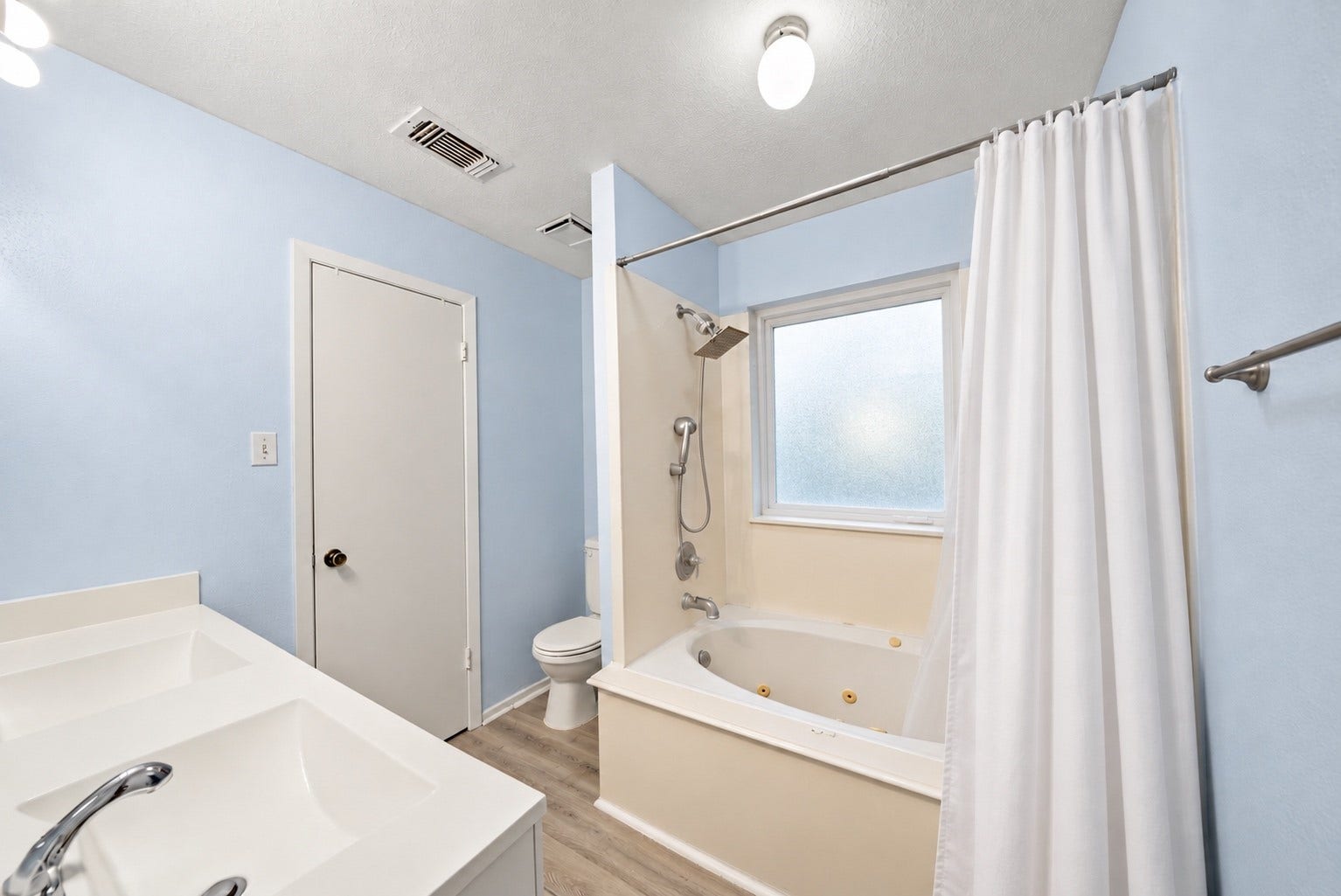Off the Primary Bedroom, the private Primary Bath has a tub with built-in jets and a handheld shower head. A frosted window over the tub lets daylight in while keeping privacy, and a white curtain hangs from the rod. The vanity has a wide basin with counter space, and the toilet sits beside the tub. A towel bar is mounted on the wall. Wood-look flooring runs through the bath, and the light blue walls match the bedroom. The jetted tub is a nice way to end a long workday!