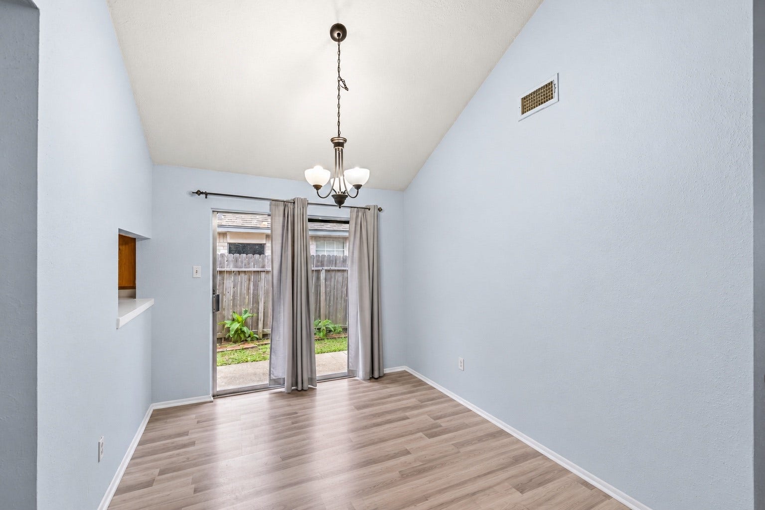 This empty angle shows the sliding glass door to the patio, the wall opening into the Kitchen on the left and a high wall vent under the vaulted ceiling. The light fixture is already hung over the table spot. Curtains on the slider give privacy at night. During the day, light from the backyard comes through the glass and across the main living area. The fenced yard is just a few steps past the patio, and the Kitchen doorway sits just to the left of this space.