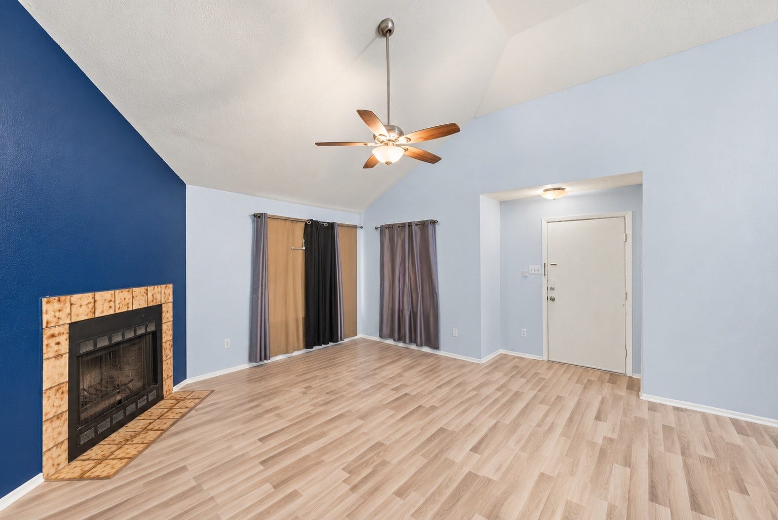 Without furniture, this view takes in the fireplace corner, both windows and the Foyer by the front door. No wall separates the Living Room from the Dining Area, and the whole front half of the home works as one open space. Wood-look floors and white baseboards carry through the whole area. The navy accent wall adds color behind the tile fireplace while the other walls stay light. A ceiling light in the Foyer and the fan light in the Living Room give good overhead light after dark!