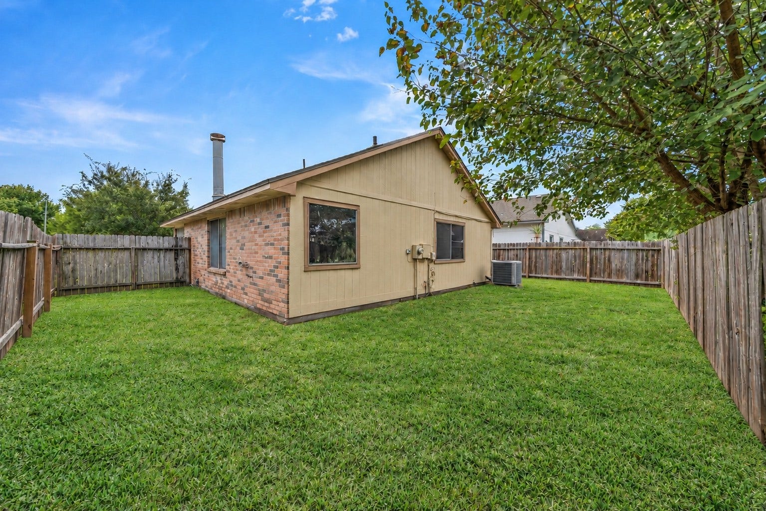 Toward the back fence, the yard opens into a wide stretch of grass behind the house, with a large tree shading the right side. From here you can see the brick and siding exterior and the chimney, with the air conditioning unit along the side wall. Sand volleyball, a baseball area and a playground at Tallow Wood Community Park are about a 6-minute walk. For groceries, H-E-B Vintage Park Market is about 2.6 miles away, and Walmart near Eldridge and FM 1960 is about 3.2 miles away.