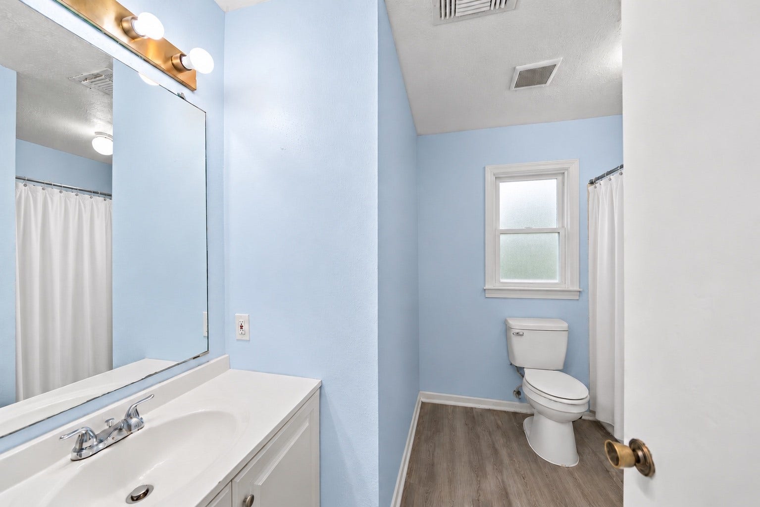 Shared by the two Secondary Bedrooms, the Hall Bath has a long vanity with counter space beside the sink, a large mirror and a bar light above. A frosted window over the toilet lets in light while keeping privacy. The shower sits behind the white curtain on the right, and the vanity cabinet is white to match the trim. Wood-look flooring runs through the bath, and the light blue walls match the bedrooms. Guests can use the Hall Bath without passing through the Primary Bedroom.