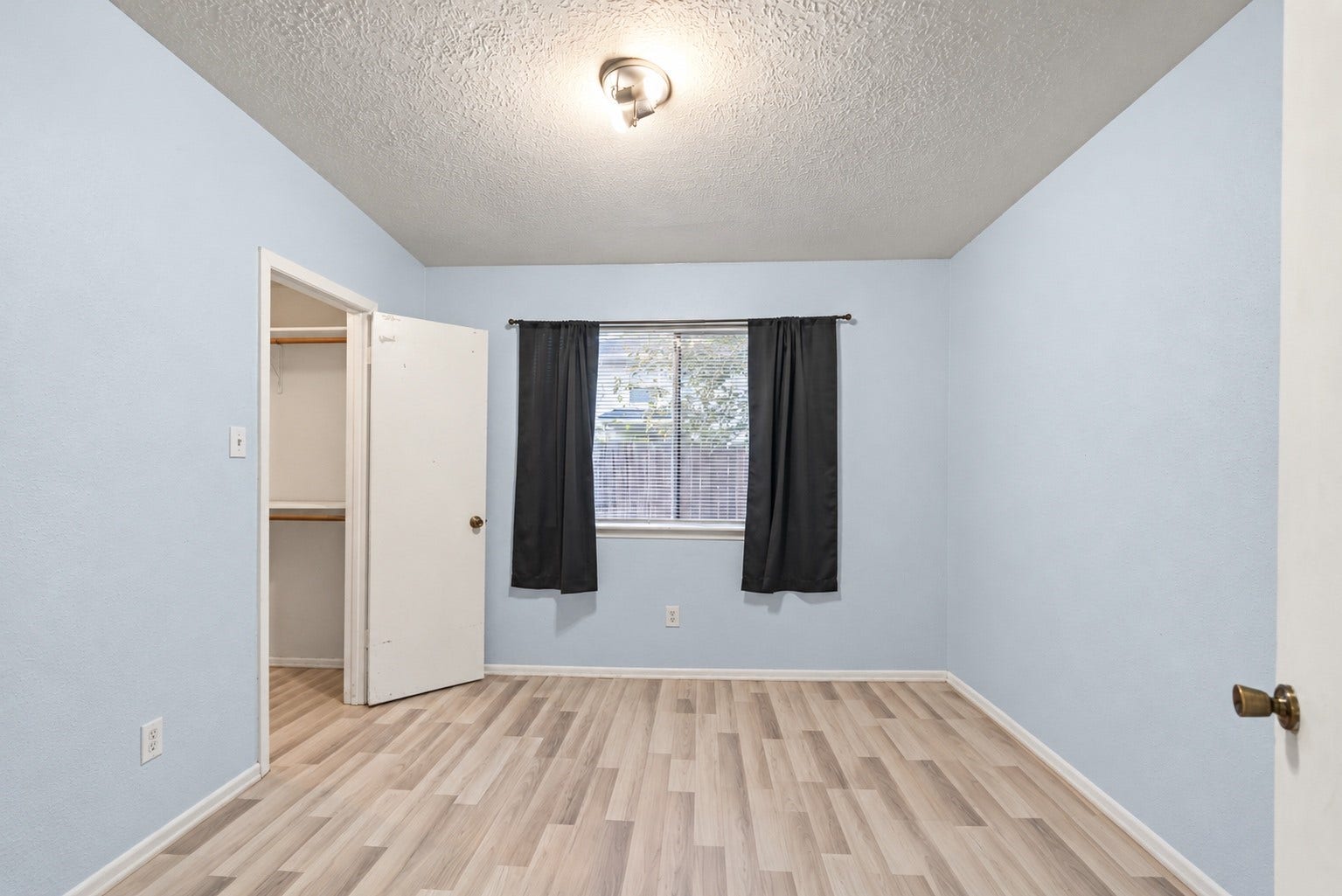 The walk-in closet (5'4" x 5'6") shows on the left in this unstaged view, with shelving and hanging space already installed. A window with curtains brings in daylight on the far wall, and a ceiling light is centered overhead. Wood-look flooring runs through, with light walls and white trim matching the rest of the home. Outlets are in place along the walls, and the door to the bedroom hall is on the right. At 10'3" x 10'2", this Secondary Bedroom has room for a full-size bed and a desk!