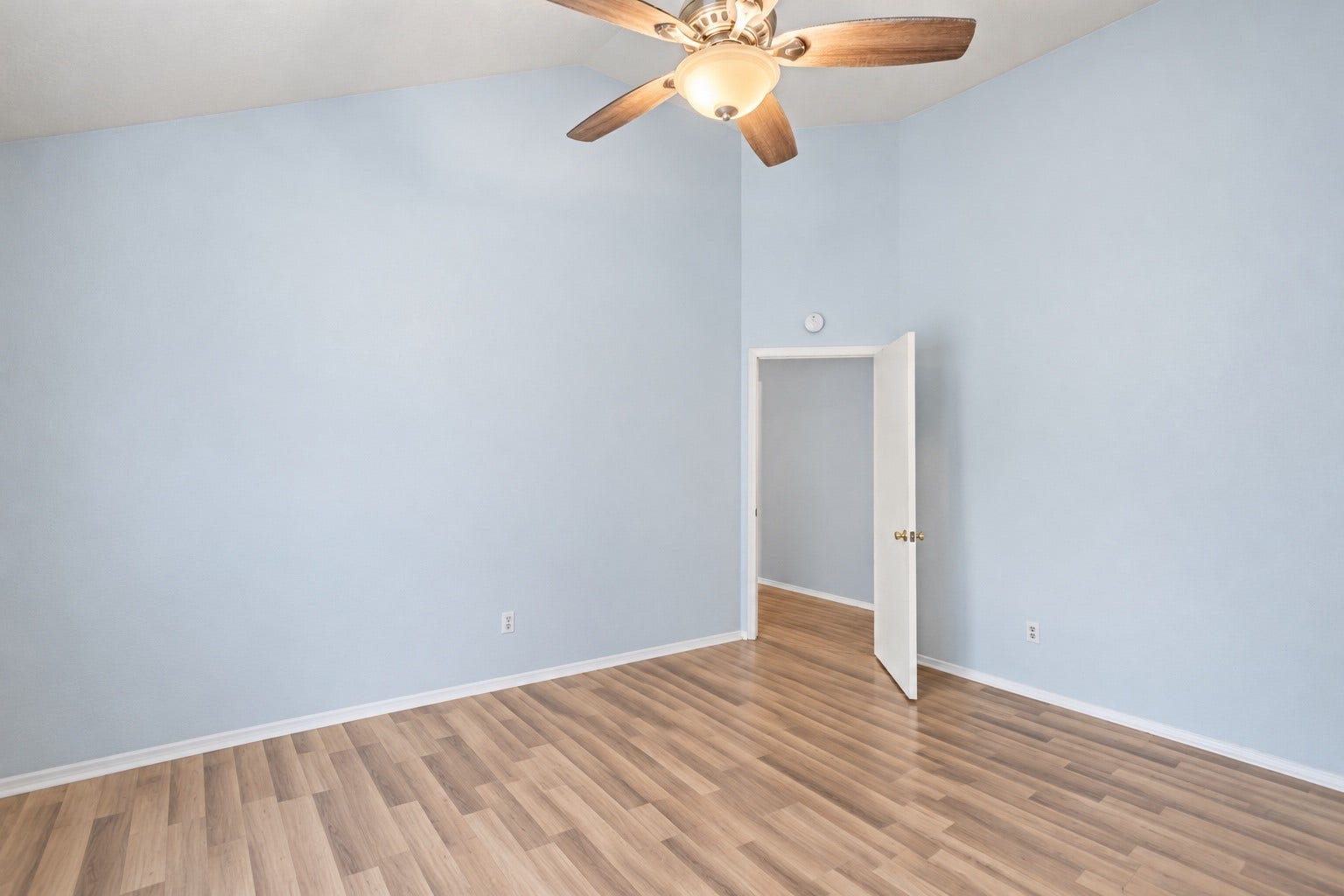 Unstaged, this angle shows the long wall for a dresser and the angled entry door from the Living Room. The Primary Bedroom sits on its own side of the home, while the secondary bedrooms are down the hall. A ceiling fan with light hangs from the vaulted ceiling. There's open floor for a reading chair or a desk, outlets sit along the walls for lamps, and wood-look floors run right to the doorway. Light blue walls with white baseboards and trim carry into the Primary Bath.