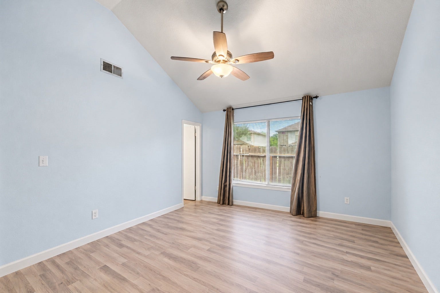 With the furniture taken out, the Primary Bedroom shows the full height of its vaulted ceiling and the big window centered on the far wall. The doorway on the left opens toward the 4'1" x 8'0" walk-in closet and the Primary Bath. Outlets are spaced around the room for lamps and electronics. Light blue walls and white baseboards carry through, and the ceiling fan helps keep the room comfortable in Houston summers. All three bedrooms are on one level!