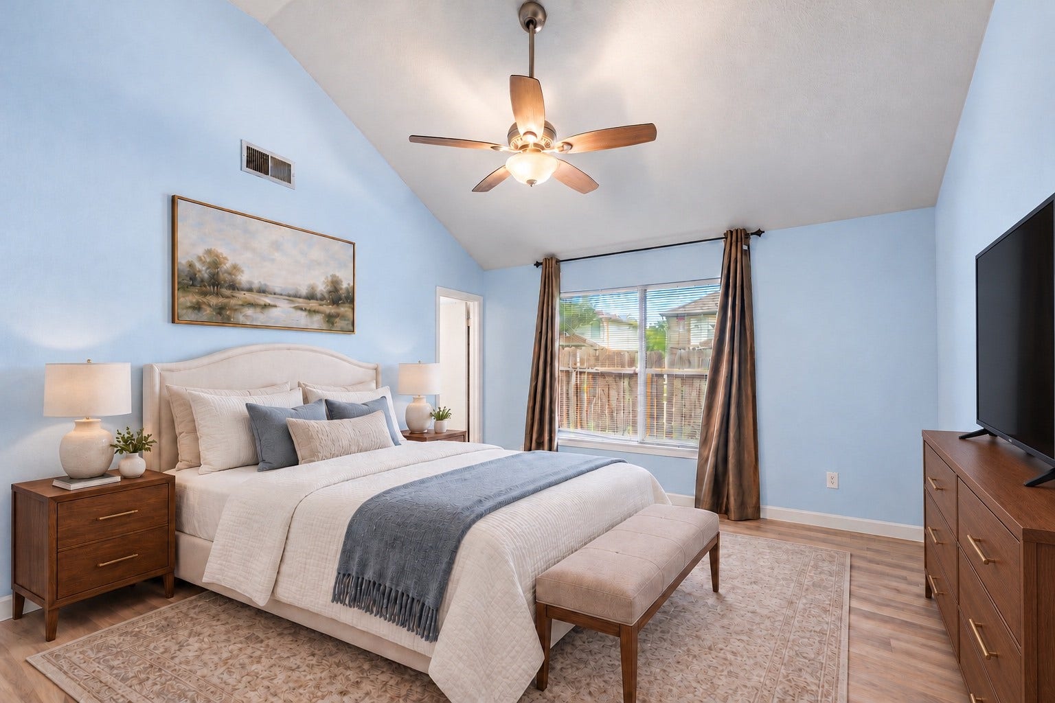 The Primary Bedroom (14'4" x 11'5") has a vaulted ceiling, a ceiling fan with light and a large window that looks out to the fenced yard. A king-size bed fits with nightstands on each side, plus a dresser on the opposite wall. Curtains are already hung on the window. The doorway on the left leads toward the walk-in closet and the private Primary Bath. Wood-look flooring continues from the Living Room, keeping the whole room carpet-free. Virtually staged.