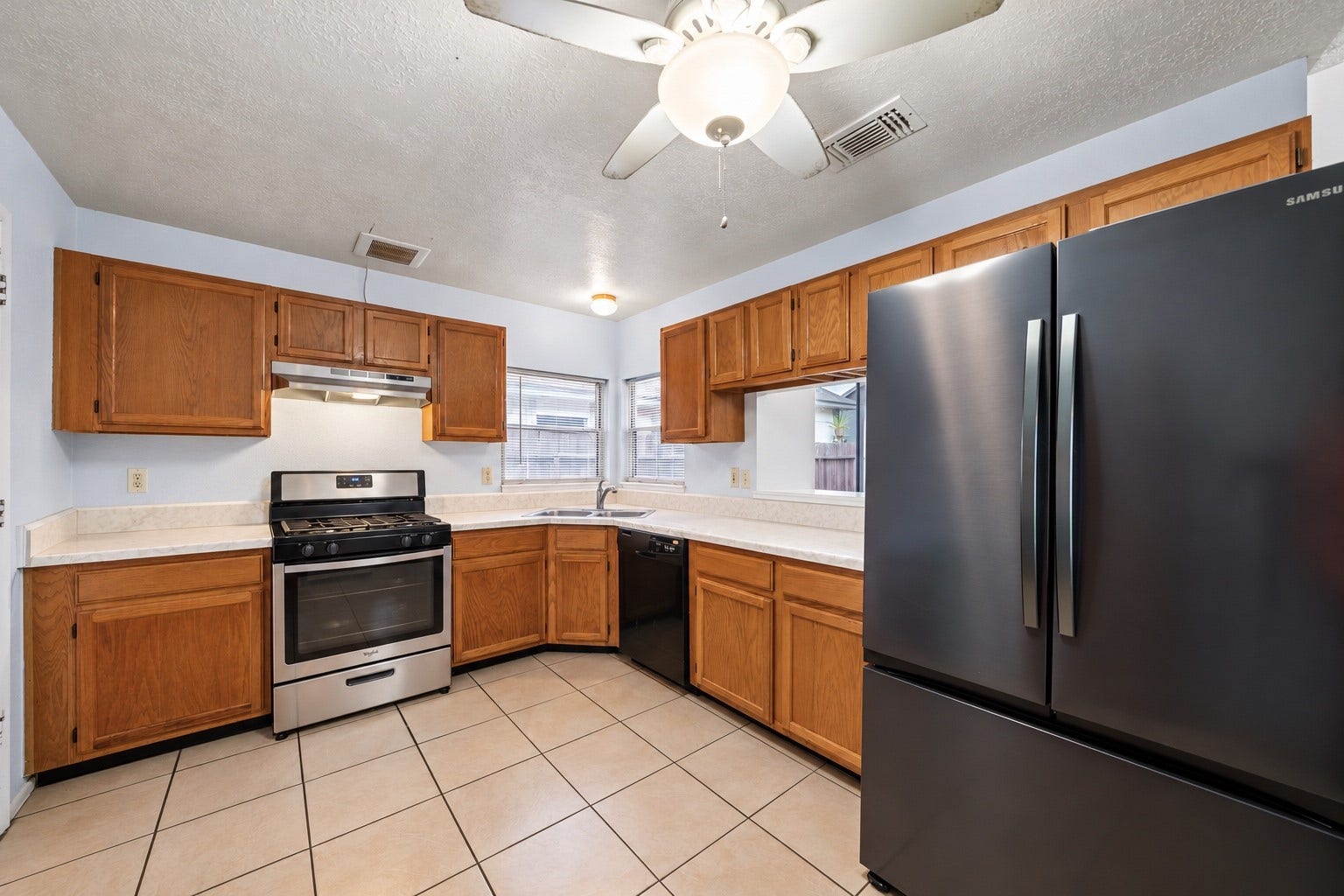 A stainless steel range sits on its own wall in the Kitchen, with a vent hood above and wood cabinets on both sides. Counters wrap around the corner to the double sink, giving prep space on each side of the stove. The black French-door refrigerator stays with the home, and a black dishwasher sits beside the sink. Windows over the sink let in daylight, and upper cabinets line both walls for dishes and pantry items. Tile flooring runs the length of the room and wipes up fast after cooking.
