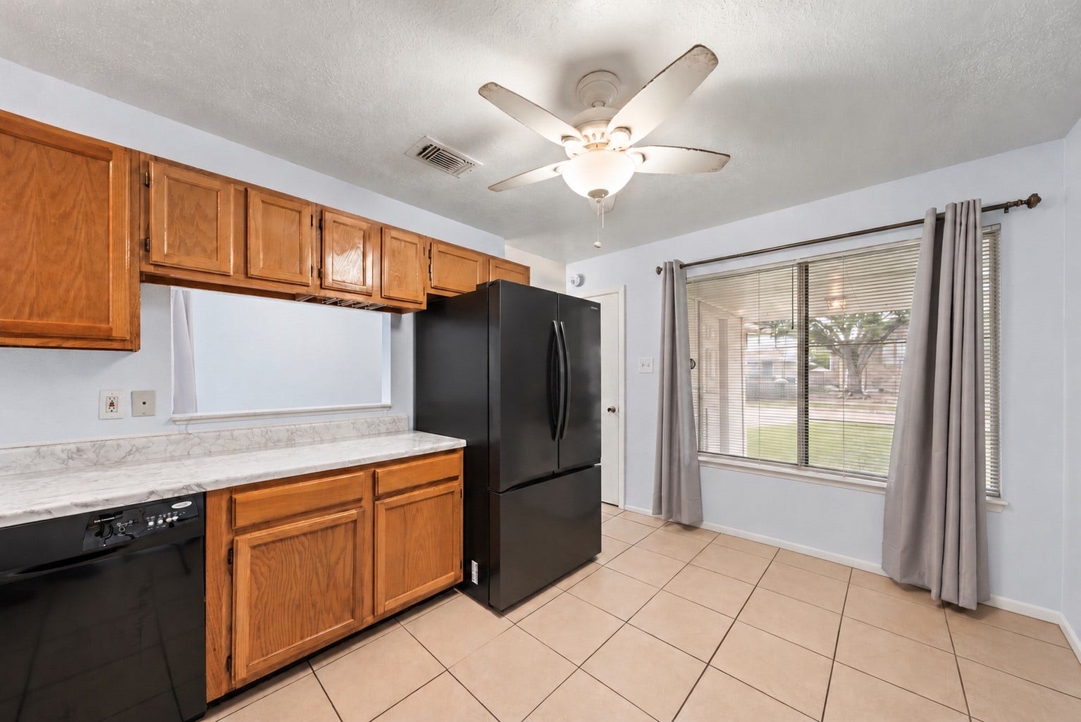 At 14'11" x 10'0", the eat-in Kitchen has room for a small table by the large curtained window. Wood cabinets and light marble-look counters run along the wall, with the black dishwasher at the end of the counter. A ceiling fan with light hangs over the room, and the tile flooring is easy to clean. Outlets along the counter leave room for small appliances, and light blue walls brighten the room. The floor plan shows direct entry from the Kitchen to the attached 2-car garage!