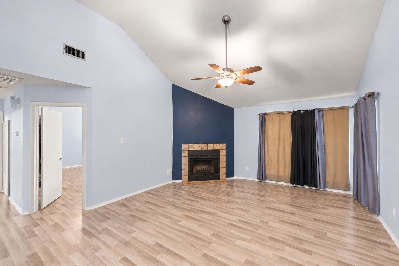 Empty, the Living Room shows its full size under the vaulted ceiling. With the fireplace tucked into the corner, the long walls stay open for a sectional or a pair of sofas. Light paint and white baseboards work with any furniture style, and the wood-look flooring means no carpet to keep up. The doorway on the left opens to the bedroom hall, with the Primary Bedroom door close by. Two windows on the far walls bring in daylight, and the ceiling fan with light is already installed.