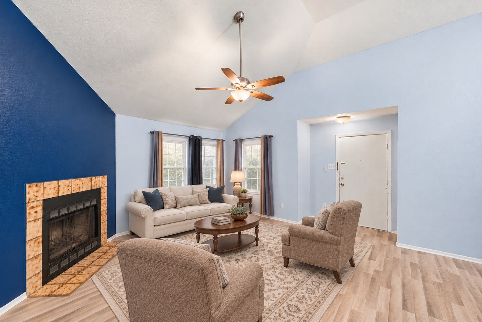 Looking back toward the front door, the Living Room has space for a sofa facing the fireplace and a pair of chairs, with open floor between the front door and the hall. Guests step from the Foyer straight into the main living space. The tile fireplace anchors the corner on the left. The vaulted ceiling with its ceiling fan runs from the fireplace toward the Dining Area. Two windows near the front door light the seating area, and white baseboards and trim finish the room. Virtually staged.
