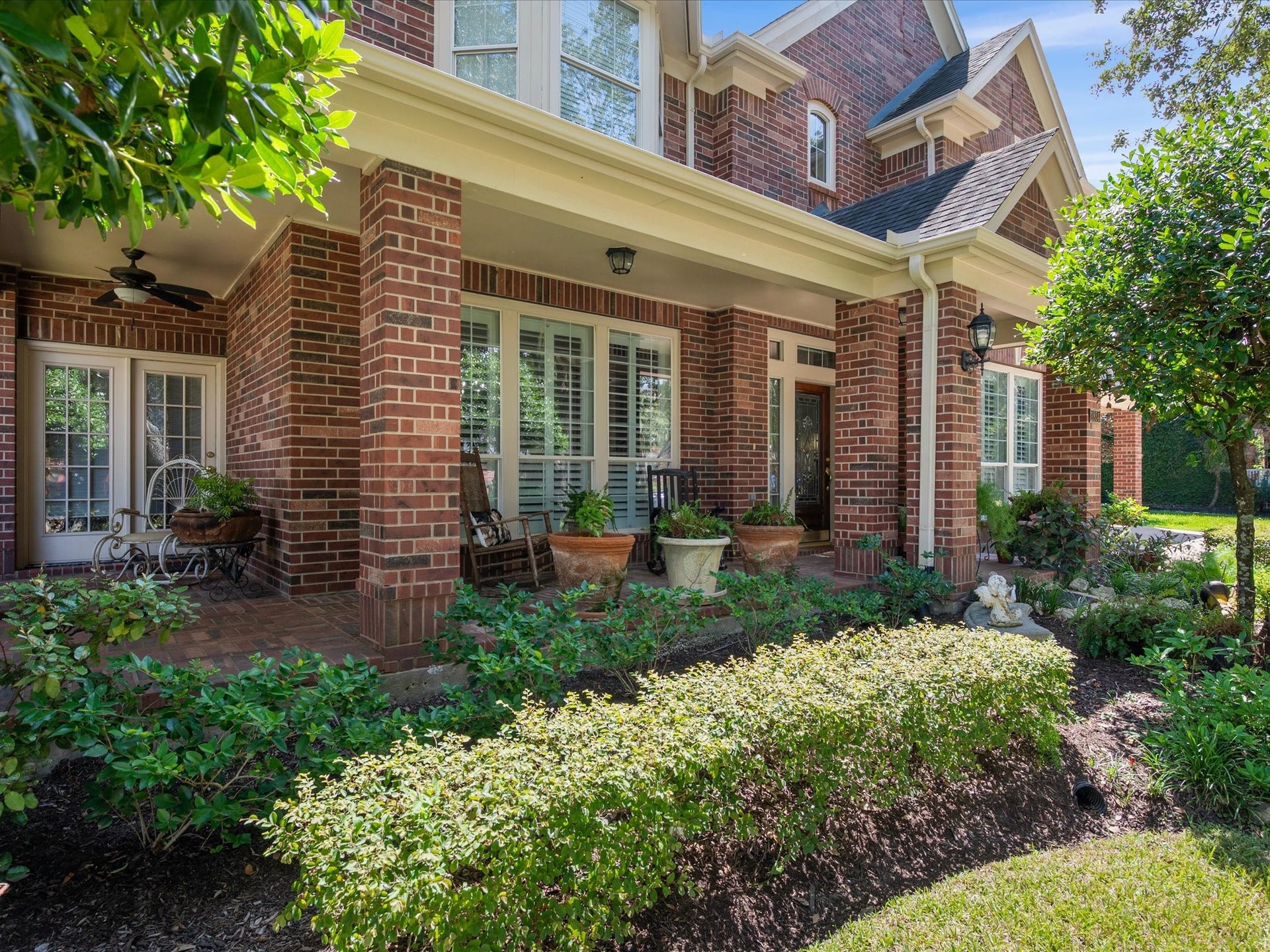 1834 Lake Quitman Drive, Richmond, TX 77406 Charming covered front porch with brick columns, a ceiling fan, and plenty of space to relax, framed by lush landscaping and potted greenery.