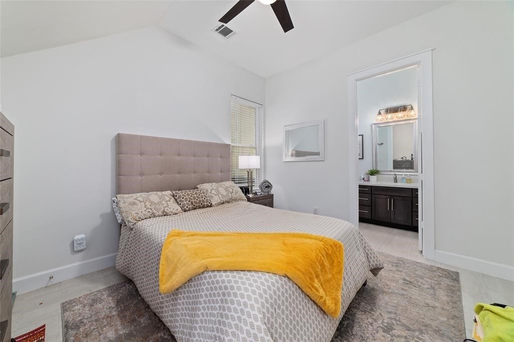 This cozy bedroom features a neutral palette with a tufted headboard and patterned bedding. It includes a ceiling fan and an en-suite bathroom with modern fixtures. The room is bright and inviting, ideal for relaxation.