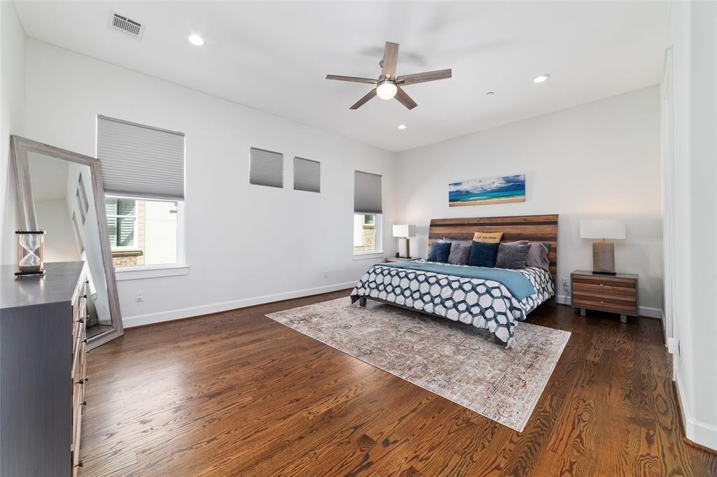 This spacious primary bedroom features hardwood floors, a large bed with modern bedding, and a ceiling fan. It includes a stylish dresser, a large mirror, and ample natural light from multiple windows. The room's neutral tones create a calming atmosphere.