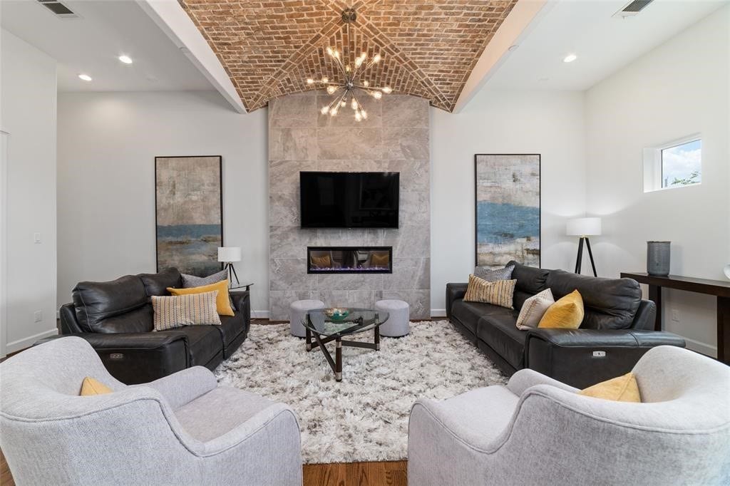 The living room features a vaulted brick ceiling, modern chandelier, and a sleek stone fireplace with a mounted TV. It includes stylish leather sofas, cozy armchairs, and a plush area rug, creating an inviting space for relaxation and entertainment.